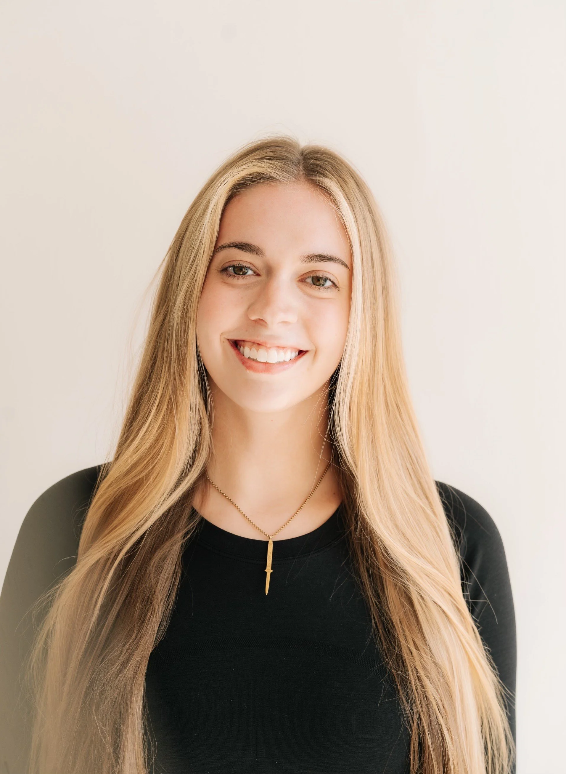 A young woman with long blonde hair, smiling, wearing a black top and a gold necklace with a small pendant, standing against a light-colored background.