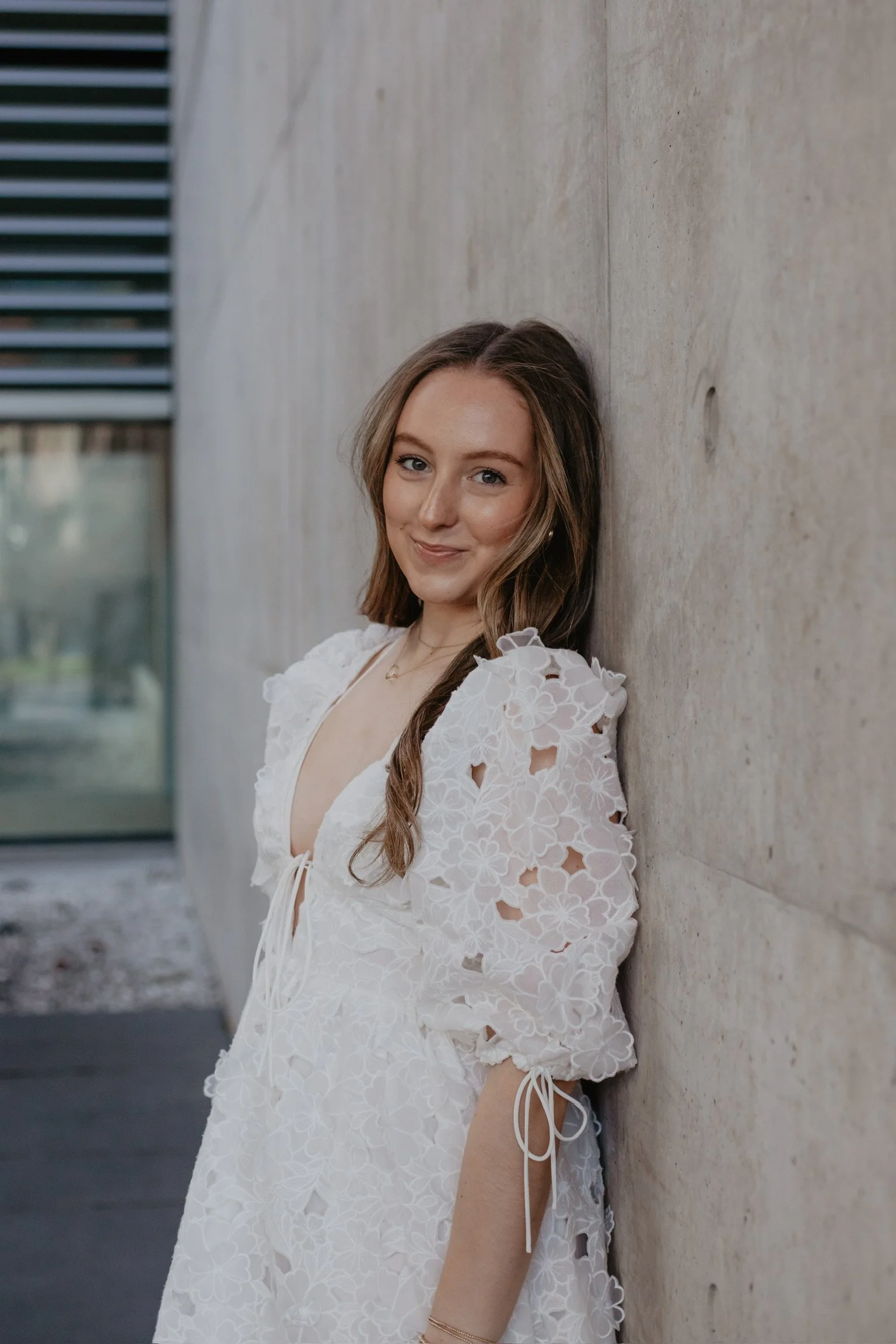 A young woman with long brown hair leaning against a concrete wall, wearing a white lace dress with floral cutouts and puff sleeves, smiling softly at the camera.