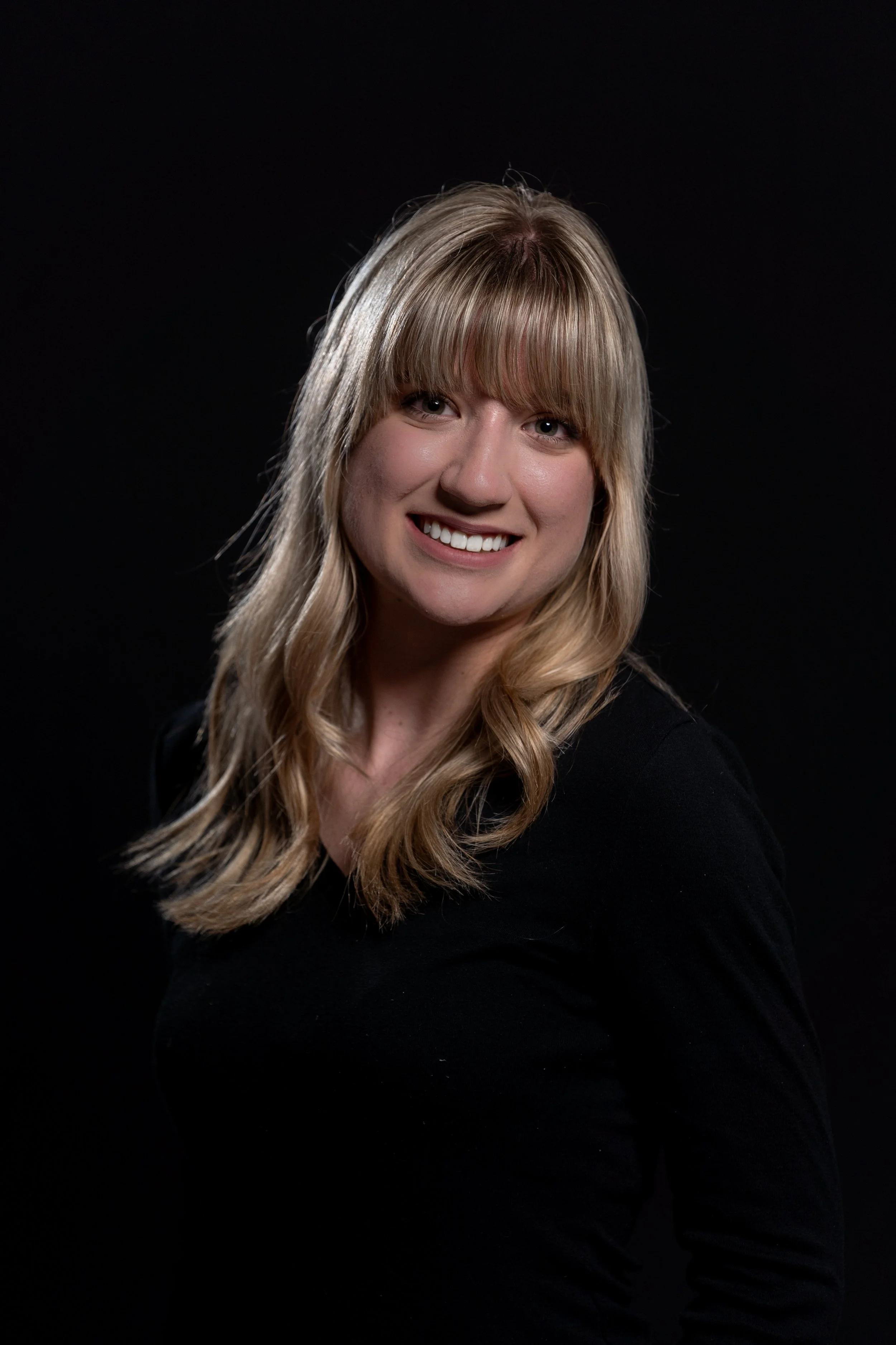A woman with long blonde hair and bangs smiling, wearing a black top, against a dark background.