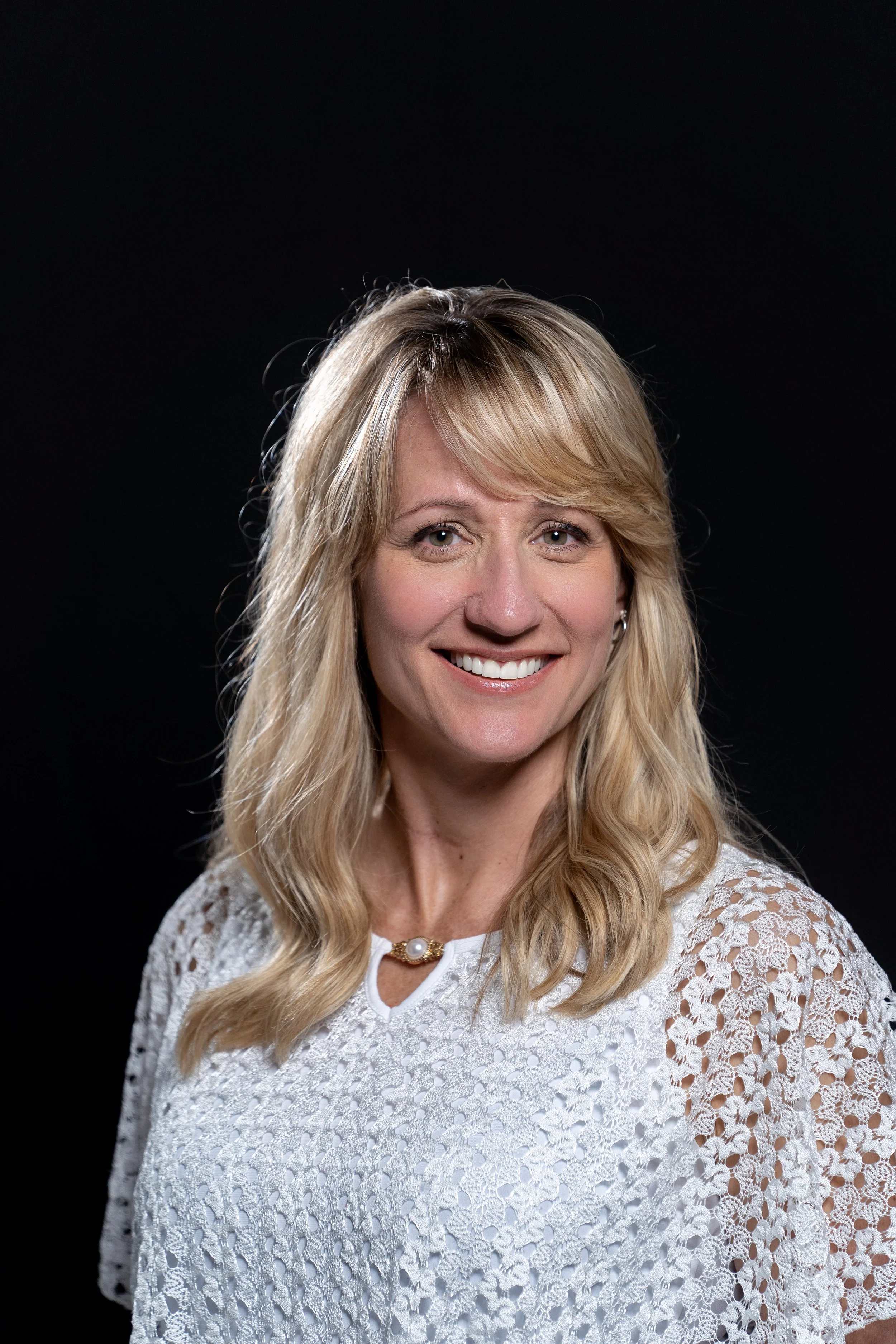 A woman with blonde hair smiling, wearing a white lace top and a pearl necklace, against a black background.