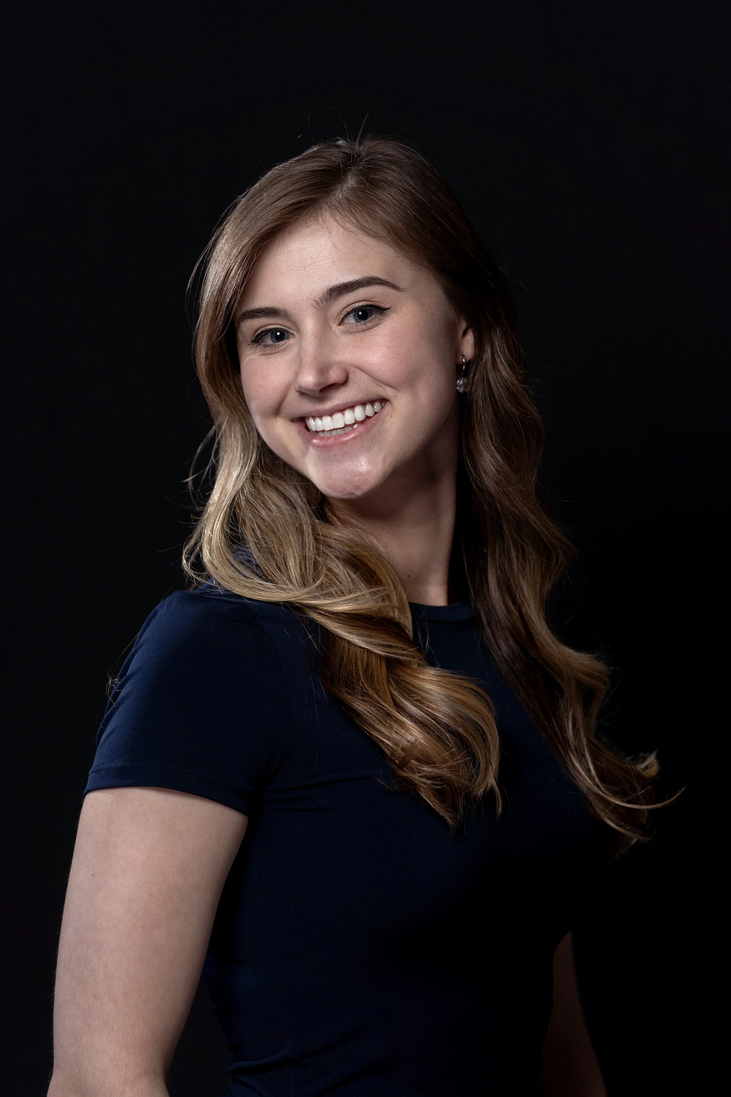 Young woman with long light brown hair, smiling in a dark background.