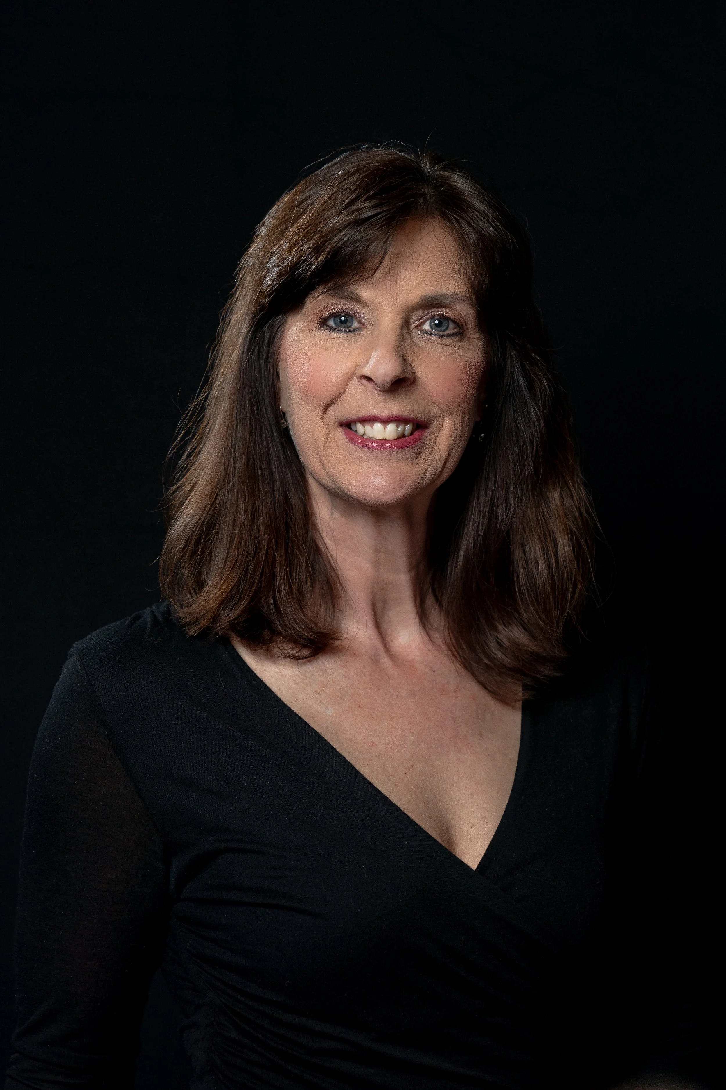 A portrait of a middle-aged woman with shoulder-length brown hair, blue eyes, and fair skin, wearing a black V-neck top, smiling against a dark background.