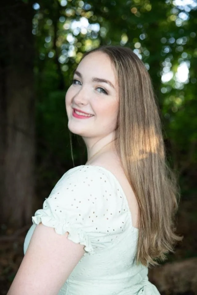 Smiling young woman with long hair standing outdoors in a forest setting.
