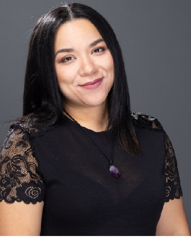 Young woman with dark hair wearing a black top with lace details and a purple pendant necklace, smiling, against a gray background.
