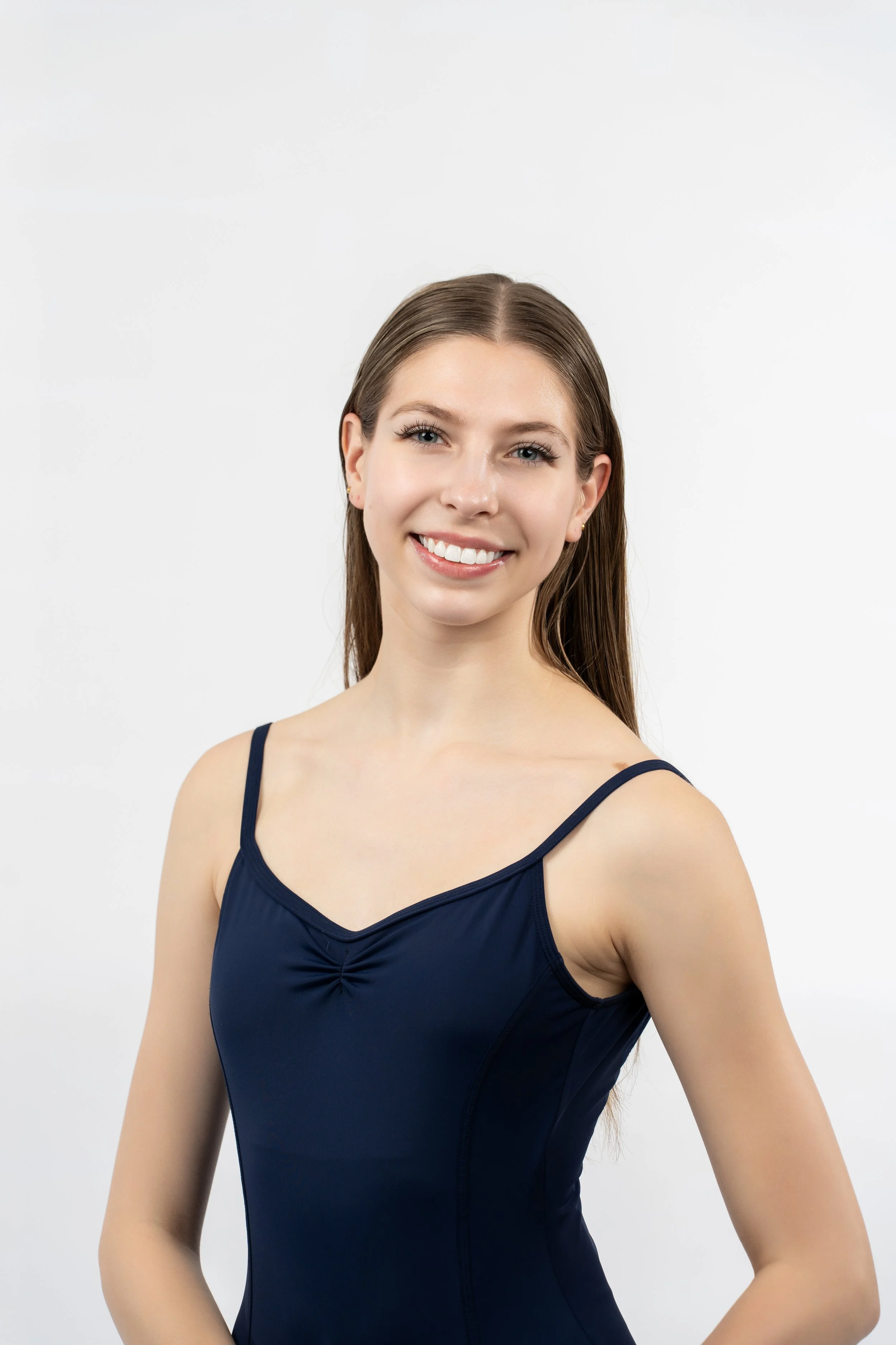 A young woman with long brown hair and a bright smile, wearing a navy blue sleeveless top with thin straps, standing against a plain white background.