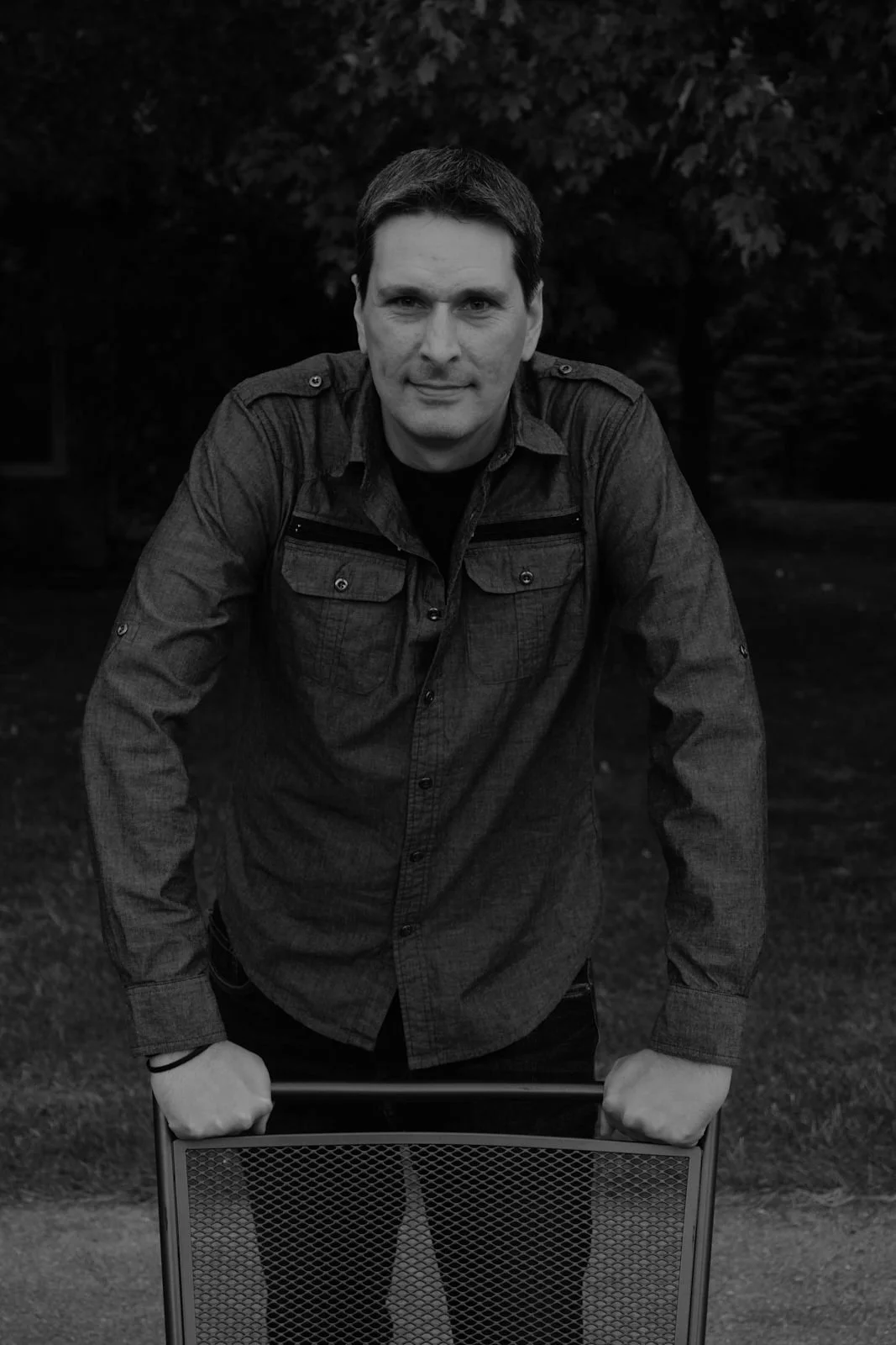 A man with dark hair wearing a dark button-up shirt, holding a chair, standing outdoors against a background of trees.
