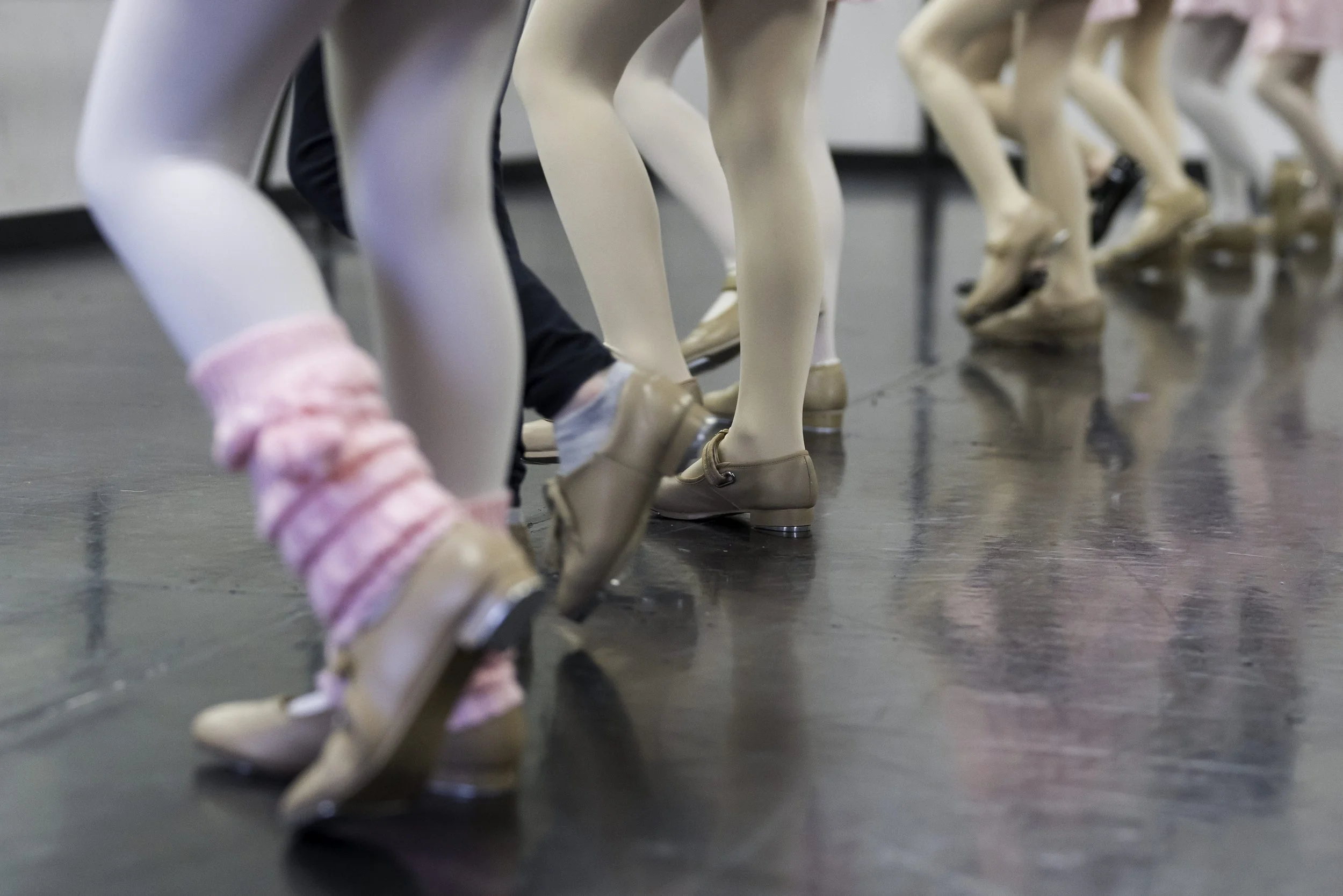 Close-up of ballet dancers' legs and feet during a dance class, wearing ballet shoes and tights, on a shiny dance studio floor.