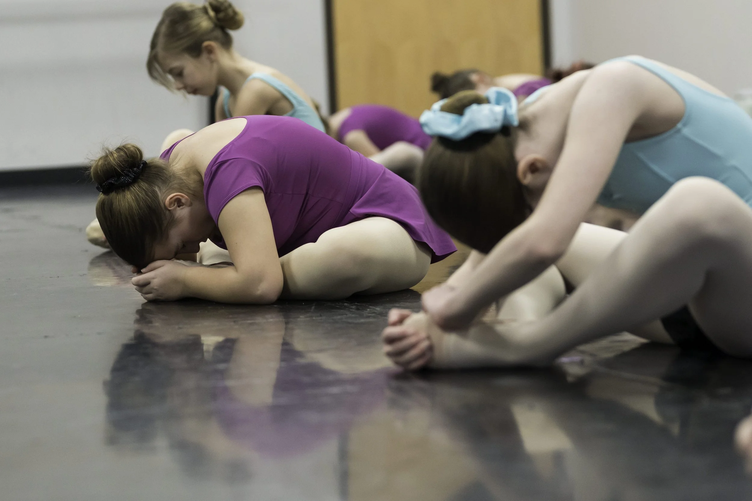 Ballet dancers practicing stretches on the floor, dressed in leotards and tights, with their heads bowed and hands clasped, in a dance studio.