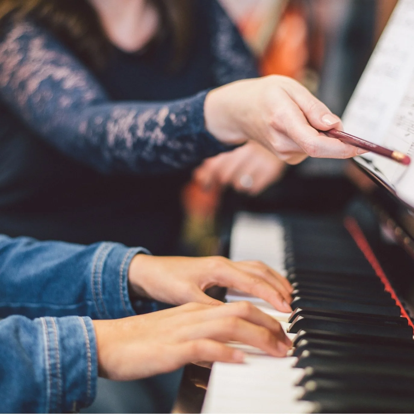 Two people playing the piano together with one person reading sheet music and the other hand on the piano keys.