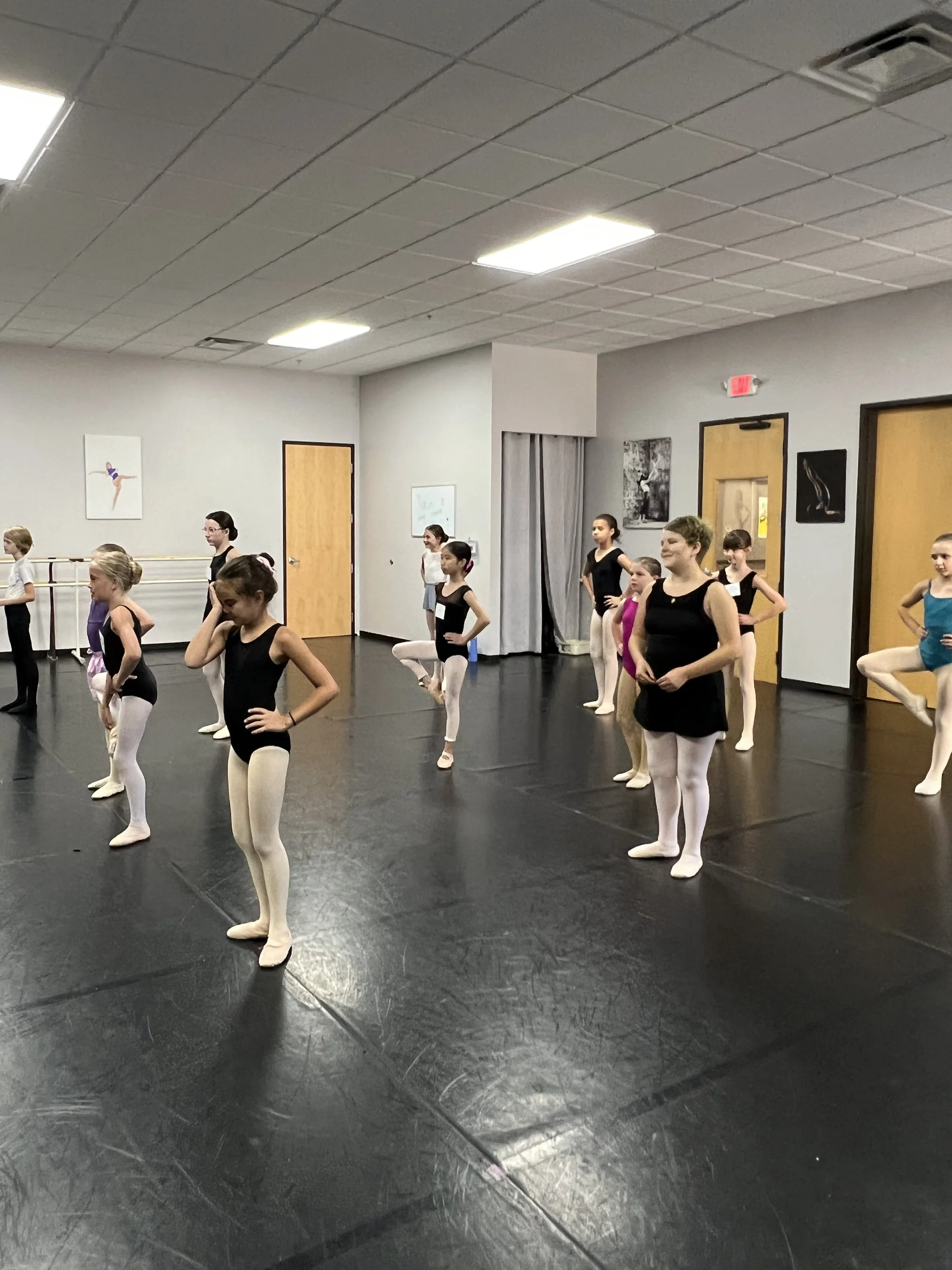 Children in ballet class practicing on a black floor with mirrors and ballet barres in the background.