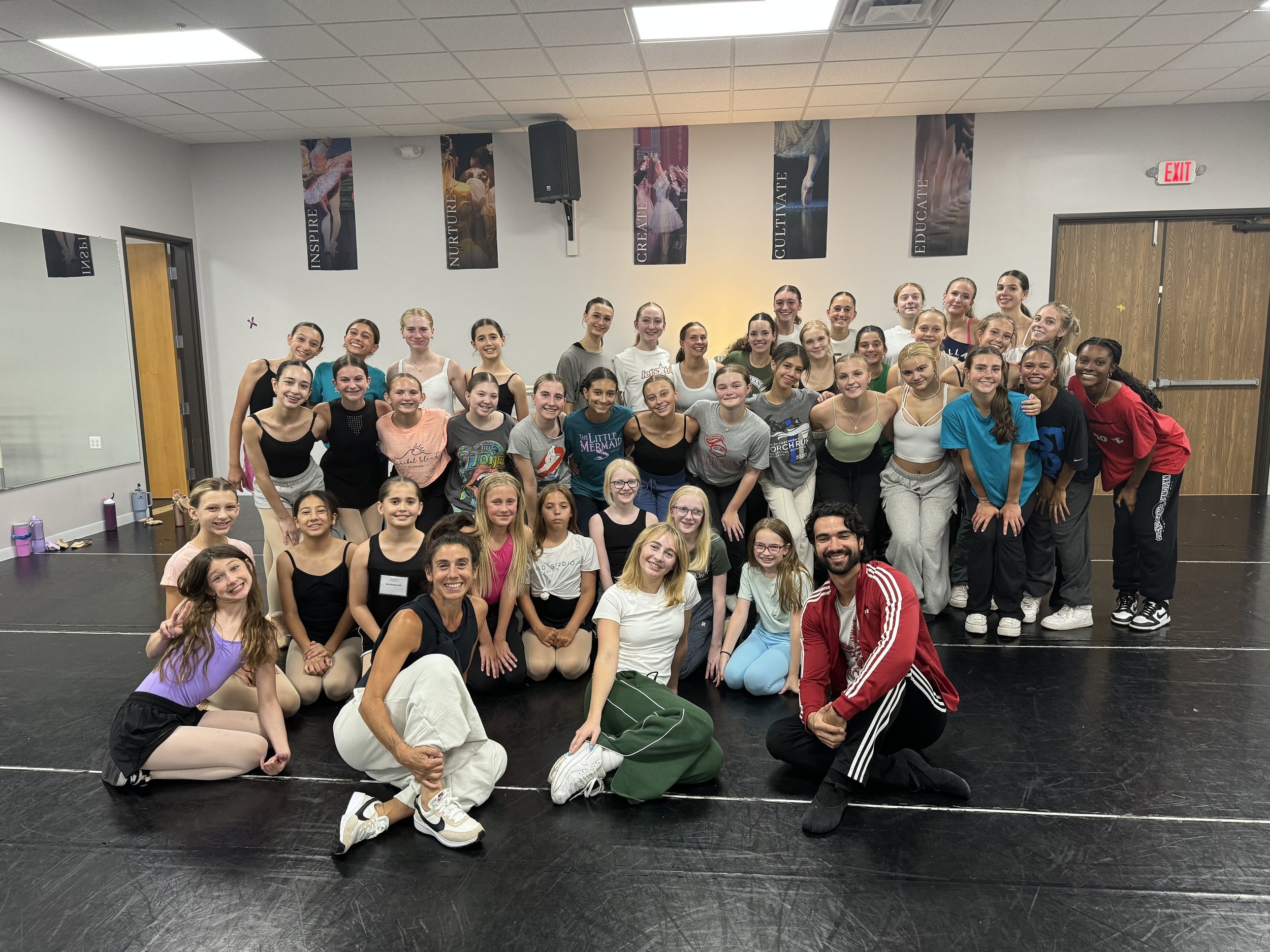 Group of young ballet dancers and two instructors posing in a dance studio with motivational banners on the wall.