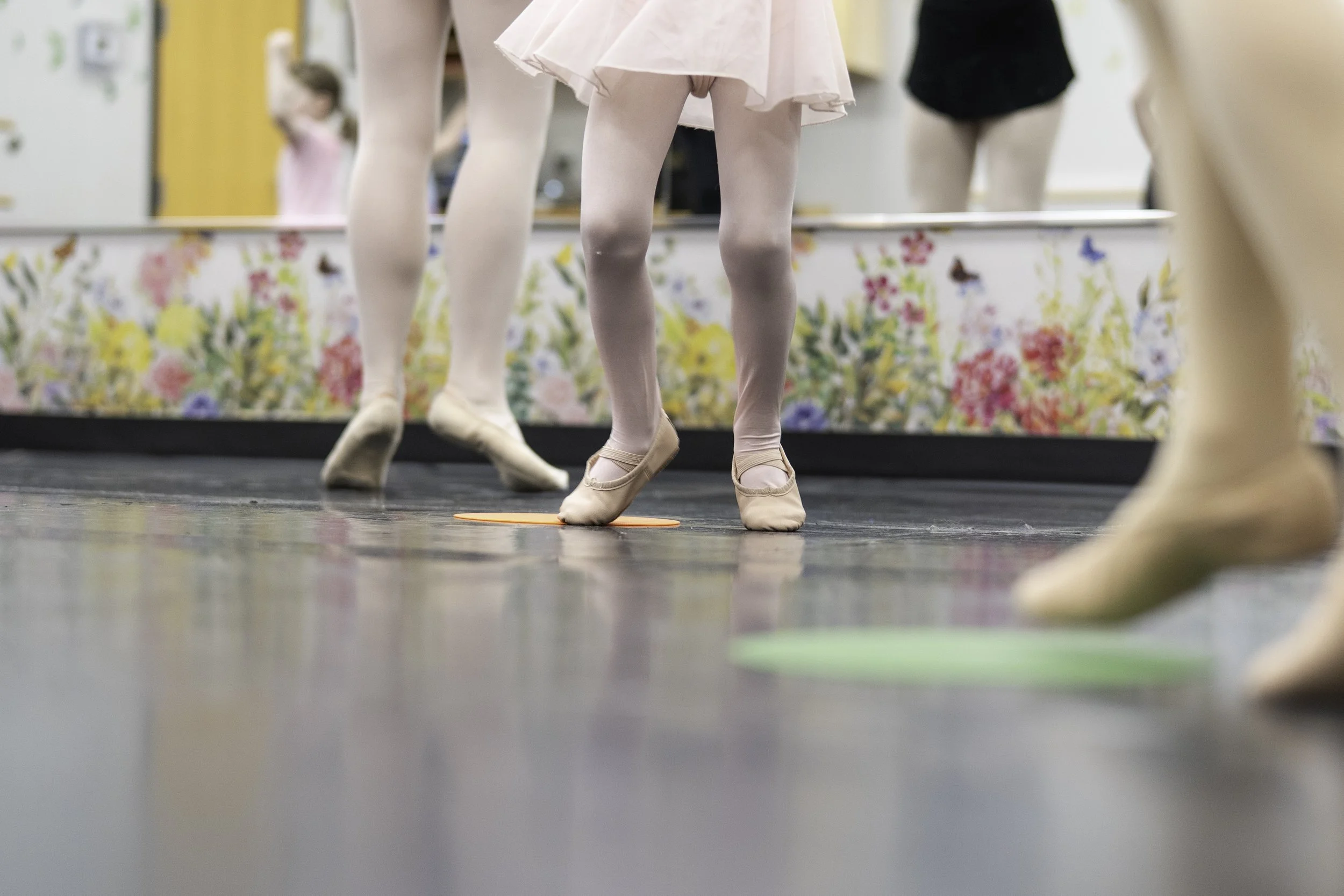 Ballet dancers practicing in a studio, focusing on their legs and feet in pink ballet slippers, with a colorful floral curtain and mirror in the background.