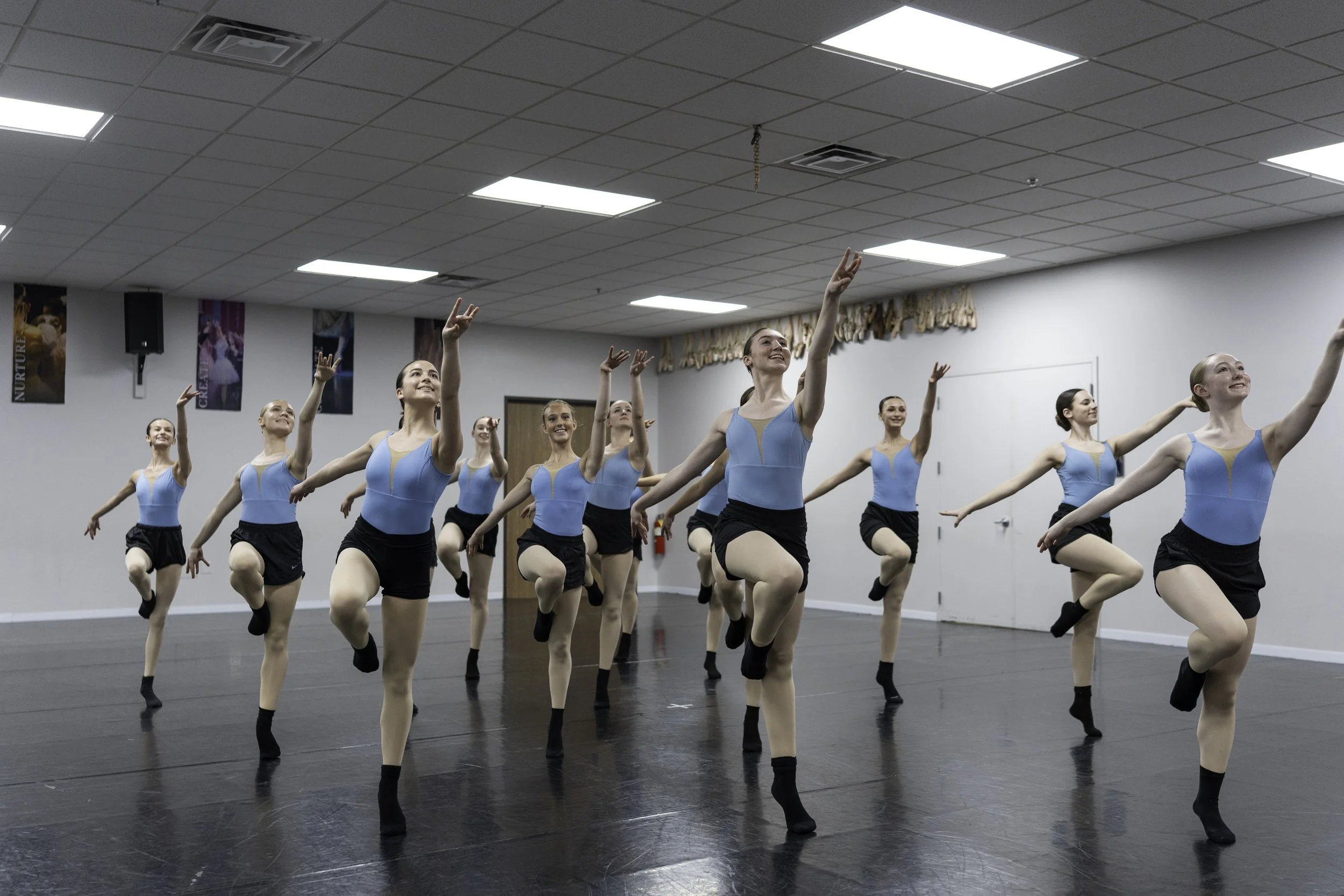 A group of ballet dancers practicing in a dance studio, wearing blue leotards and black shorts, performing a synchronized dance with one leg raised and arms extended.