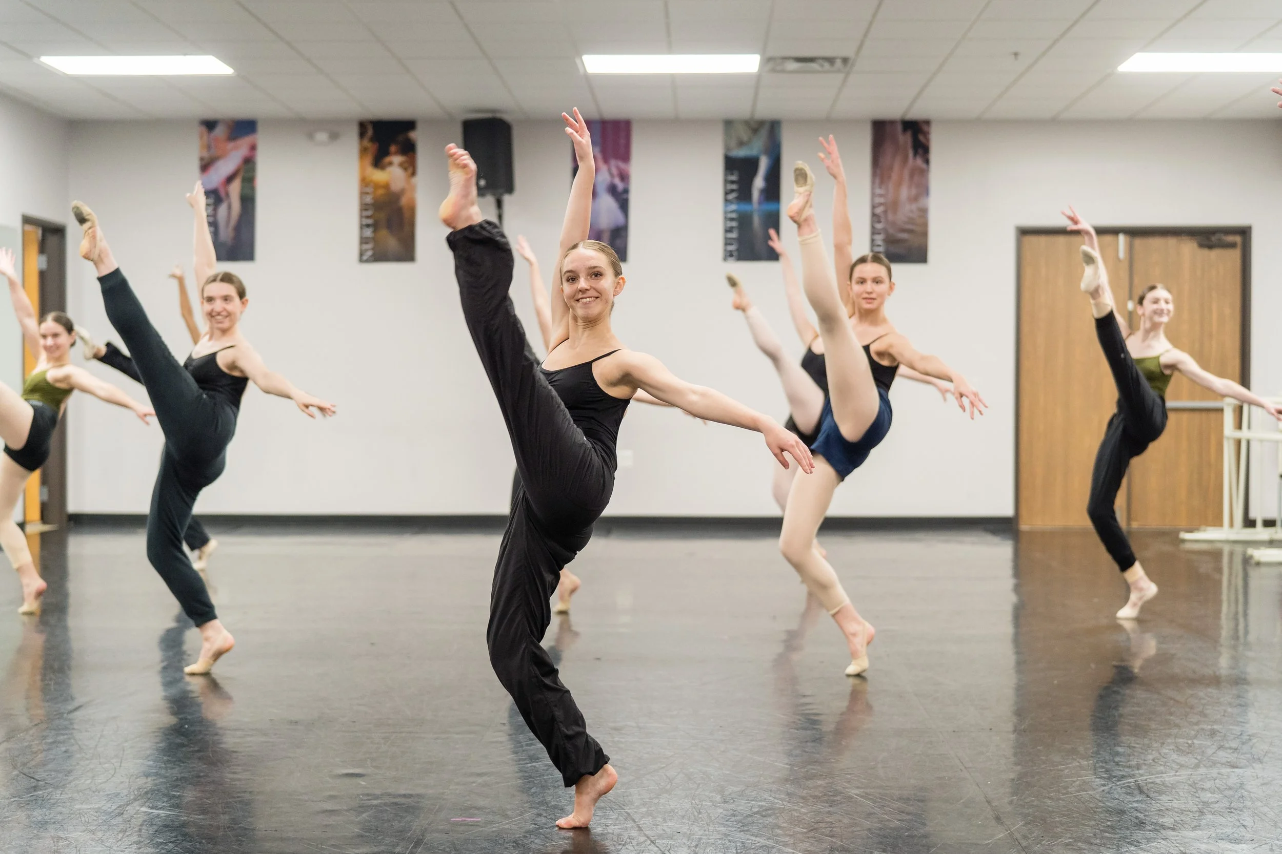 Ballet dancers practicing in a dance studio with white walls and posters, all performing a high leg extension.