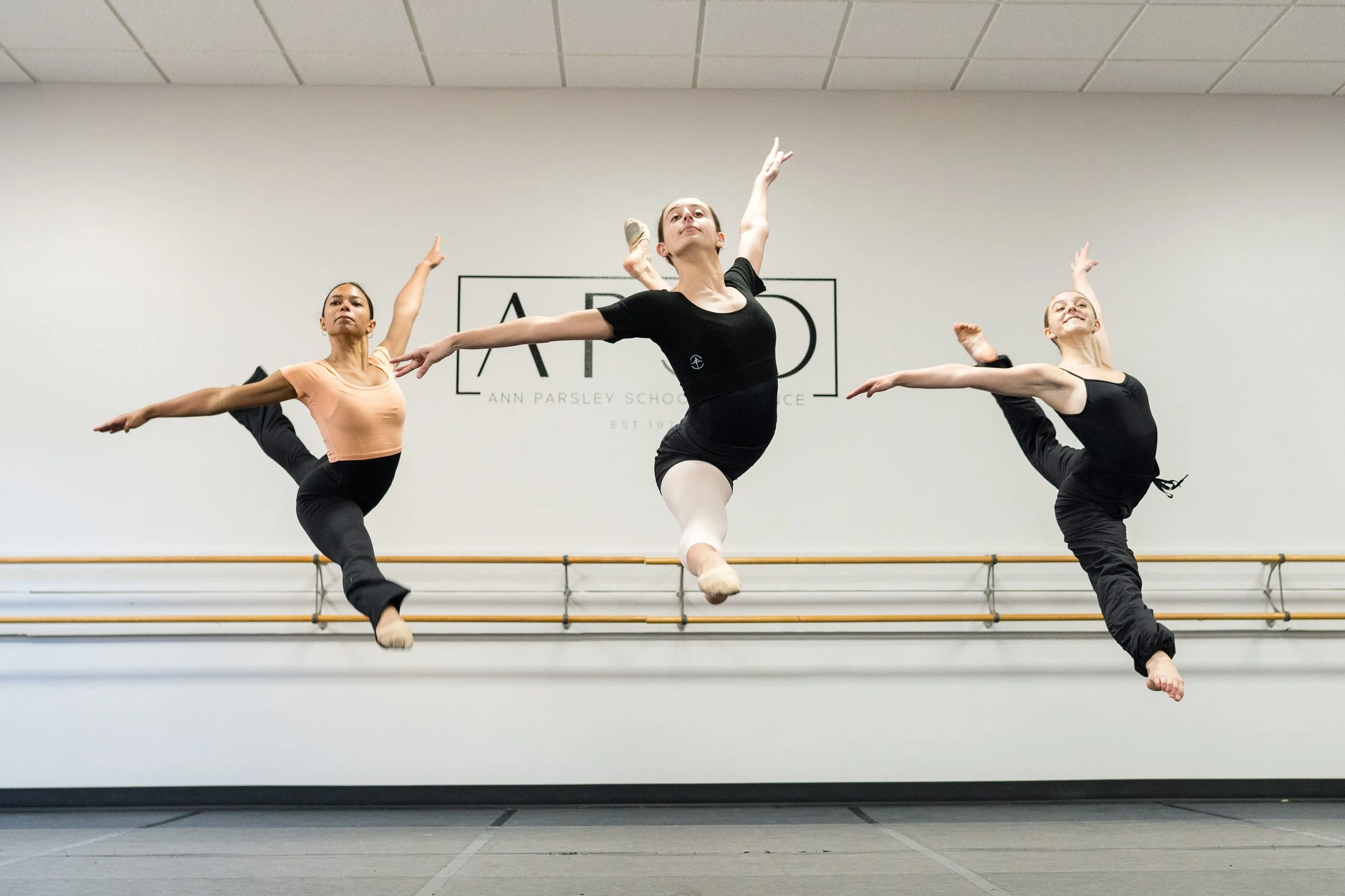Three ballet dancers mid-air during rehearsal at Ann Parsley School of Dance with a plain wall and ballet barre in the background.