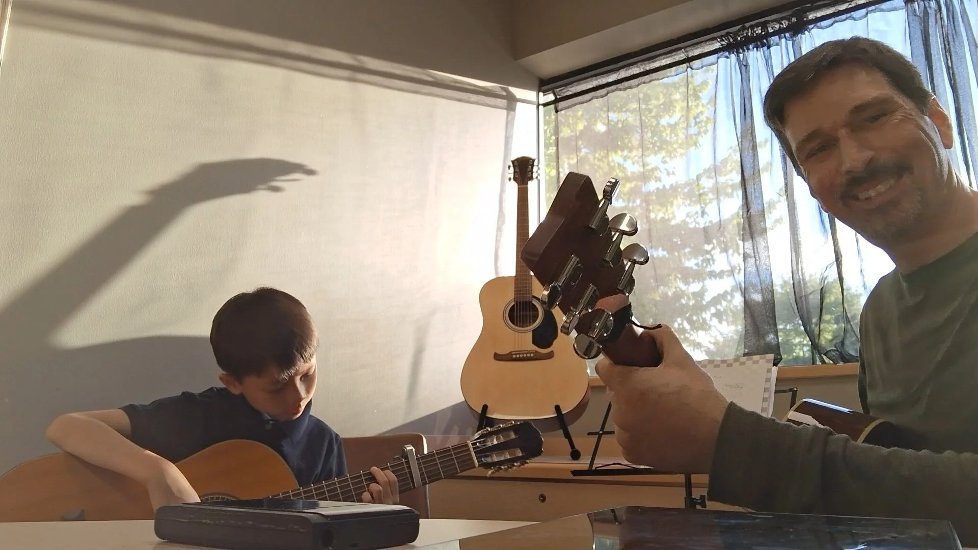 A man and a boy playing acoustic guitars indoors. The man is smiling and holding a guitar, while the boy is focused on his guitar. There is a guitar on a stand, a music stand, and a window with curtains in the background.