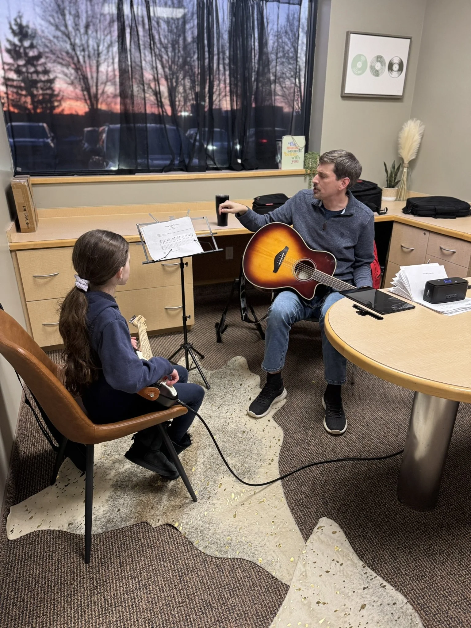 A man playing guitar and singing while a young girl listens in a small, indoor room with a window showing a sunset sky and trees outside.