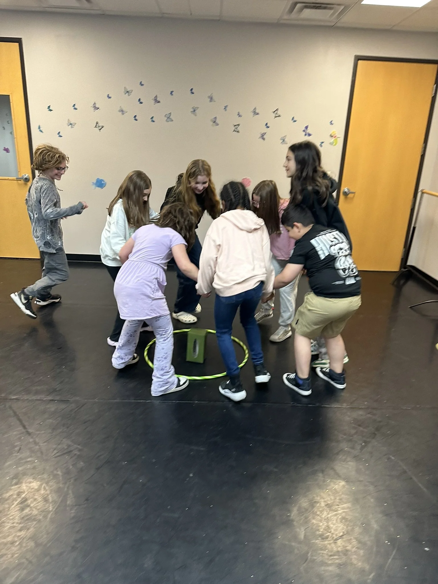 A group of children gathered in a circle playing a game with a hoop on the floor inside a room decorated with butterfly wall decals.