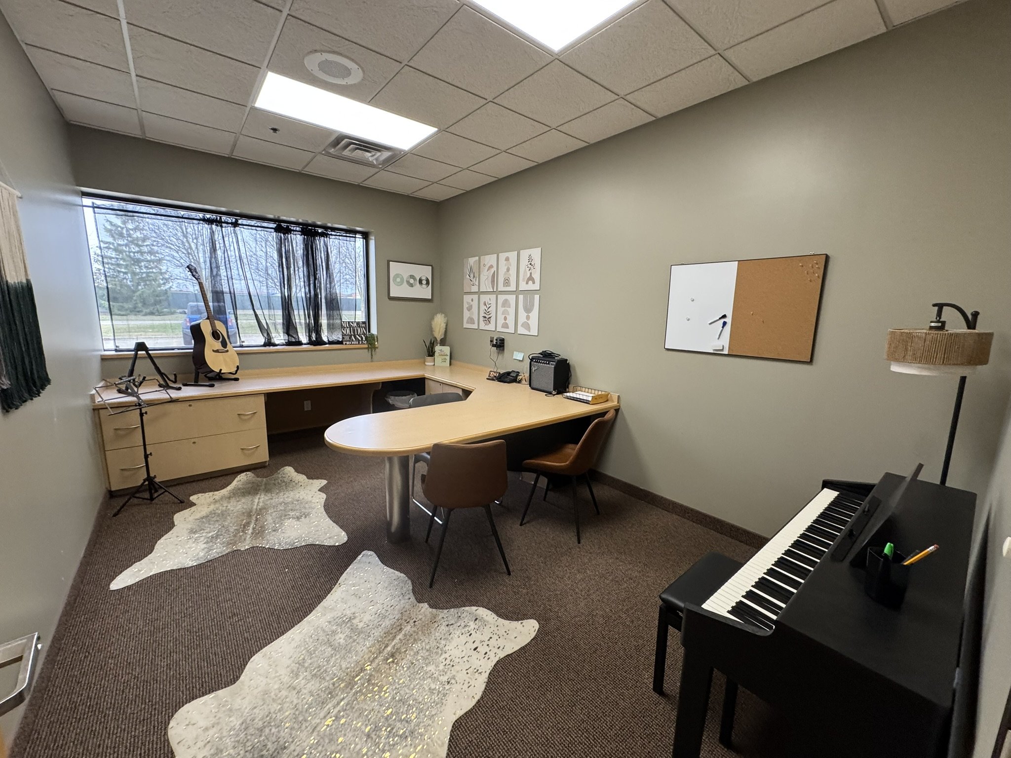 An office space with a large window, a desk with a guitar, microphone, and various office supplies, beige wall with artwork, a whiteboard, a keyboard, and a cowhide rug on the brown carpeted floor.
