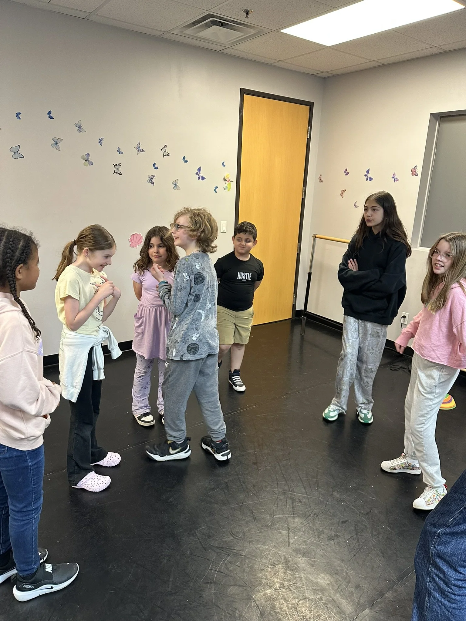 Group of children standing in a circle in a room with white walls decorated with butterfly stickers. The children are casually dressed and appear to be engaged in a conversation or activity.