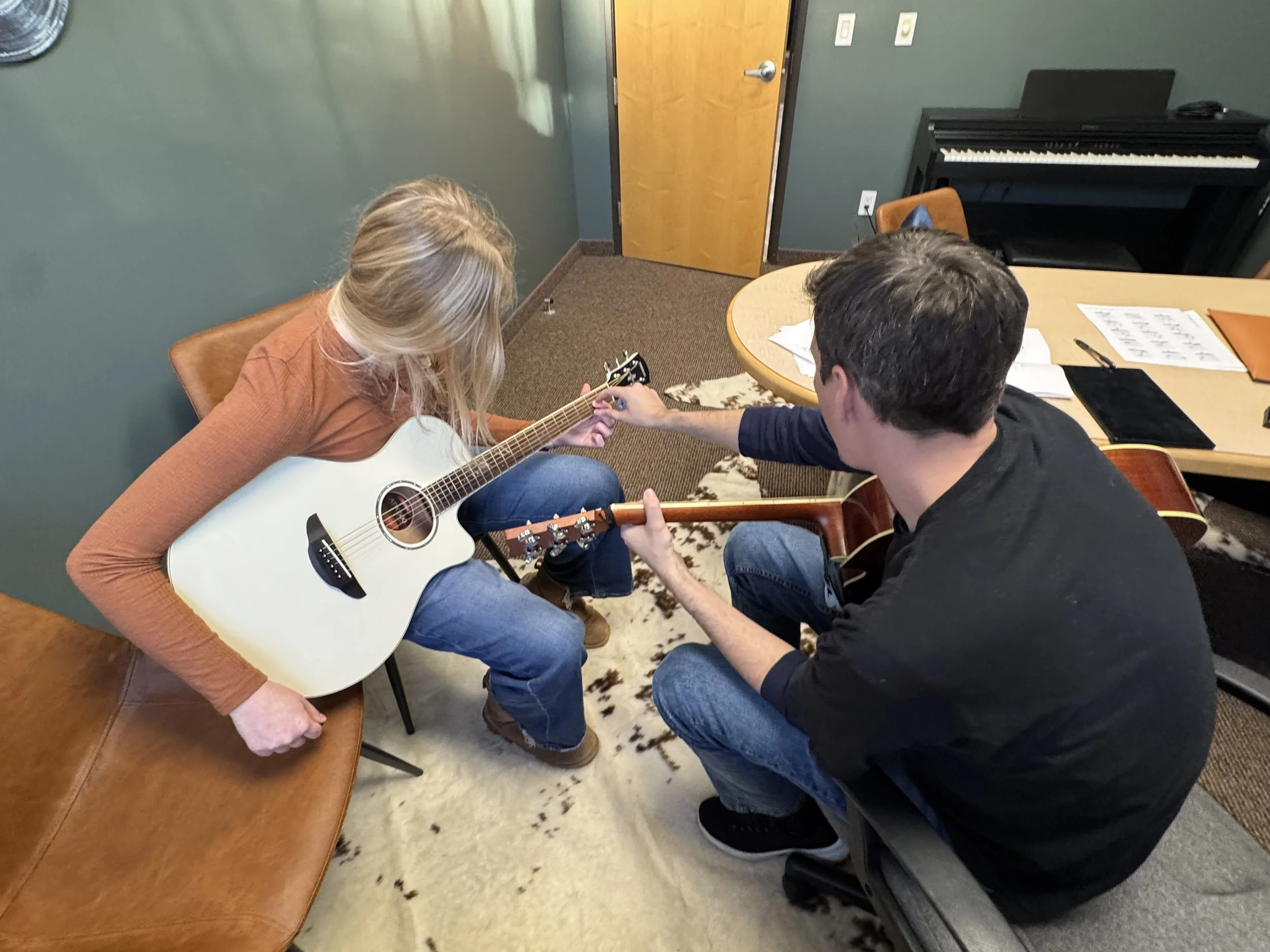Two people sitting at a table, with a woman holding a white guitar, while the man offers help or guidance, in a room with a piano and a closed door.