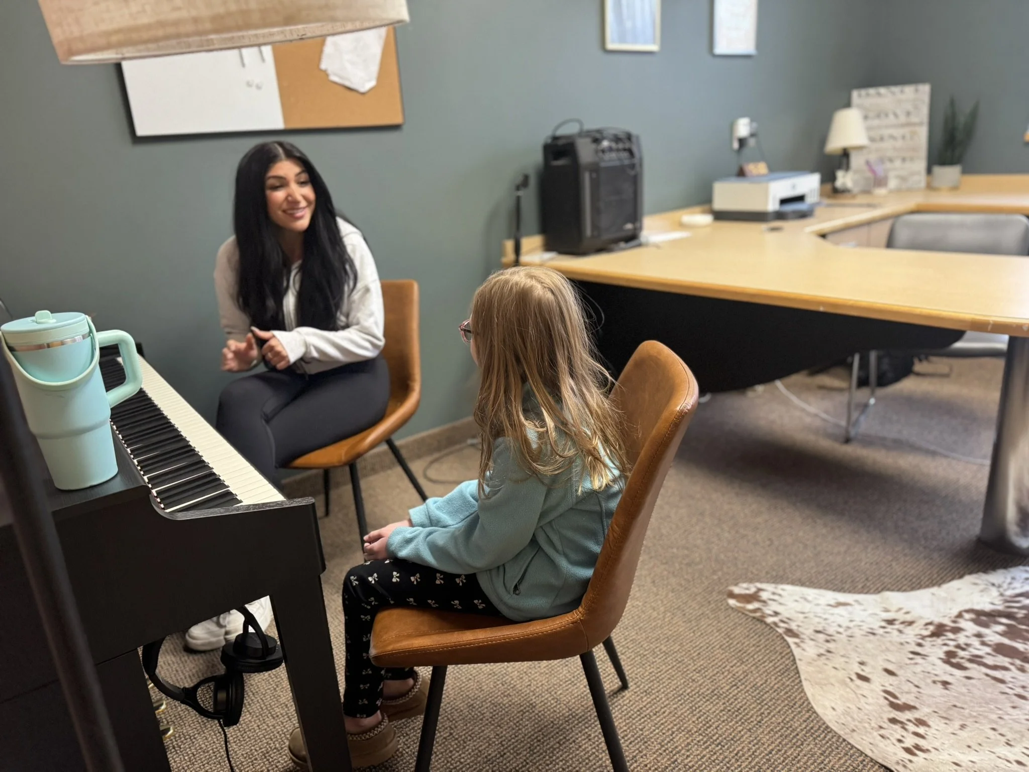 A woman with long dark hair and a white top smiling while sitting in a brown chair across from a young girl with blonde hair and glasses in a green hoodie in an office or classroom setting, near a black digital keyboard.