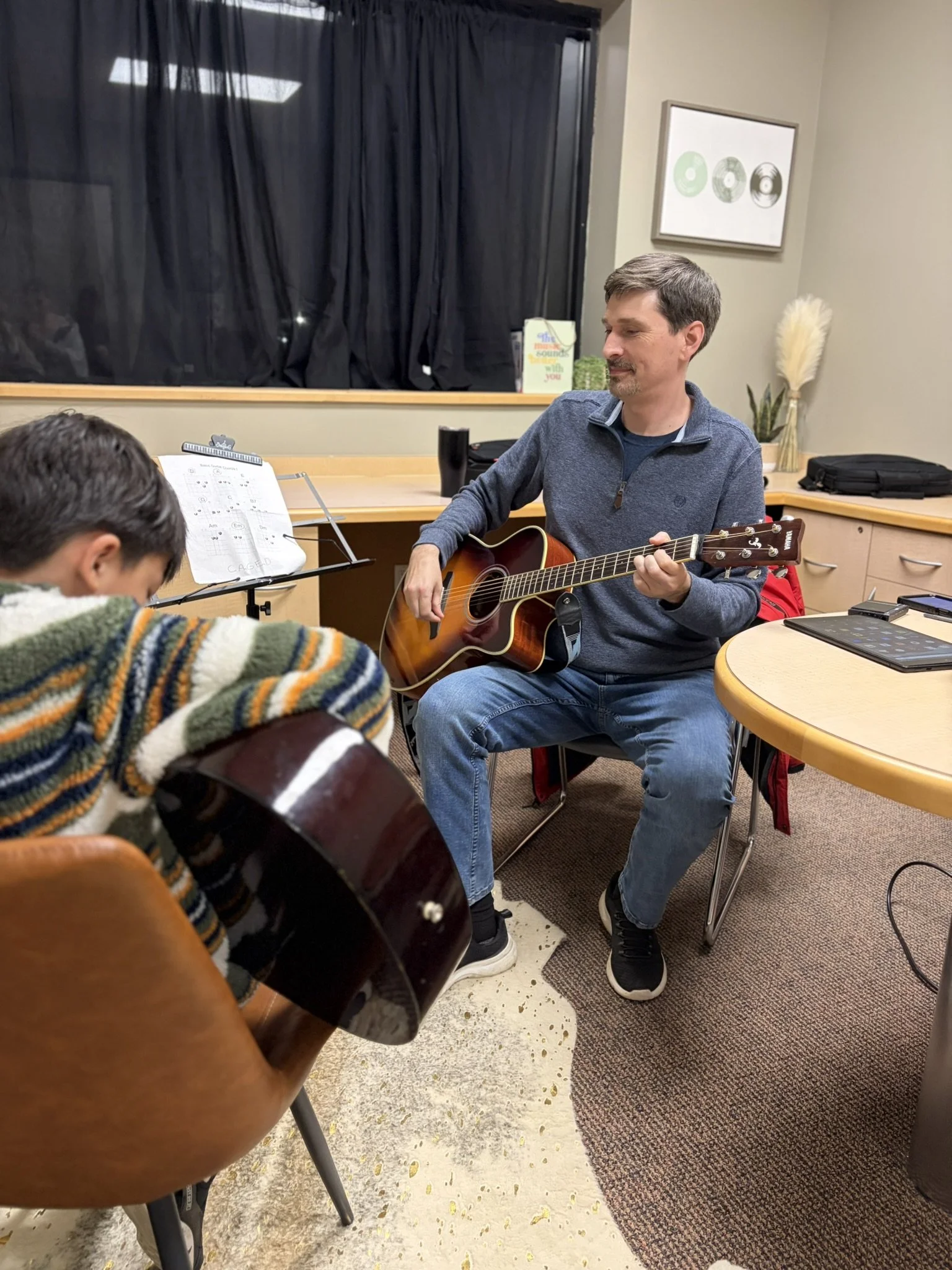 A man sitting on a chair playing an acoustic guitar in a room with beige walls and a black curtain covering a window. There are musical notes on a stand, a framed picture on the wall, and a vase with pampas grass in the background.