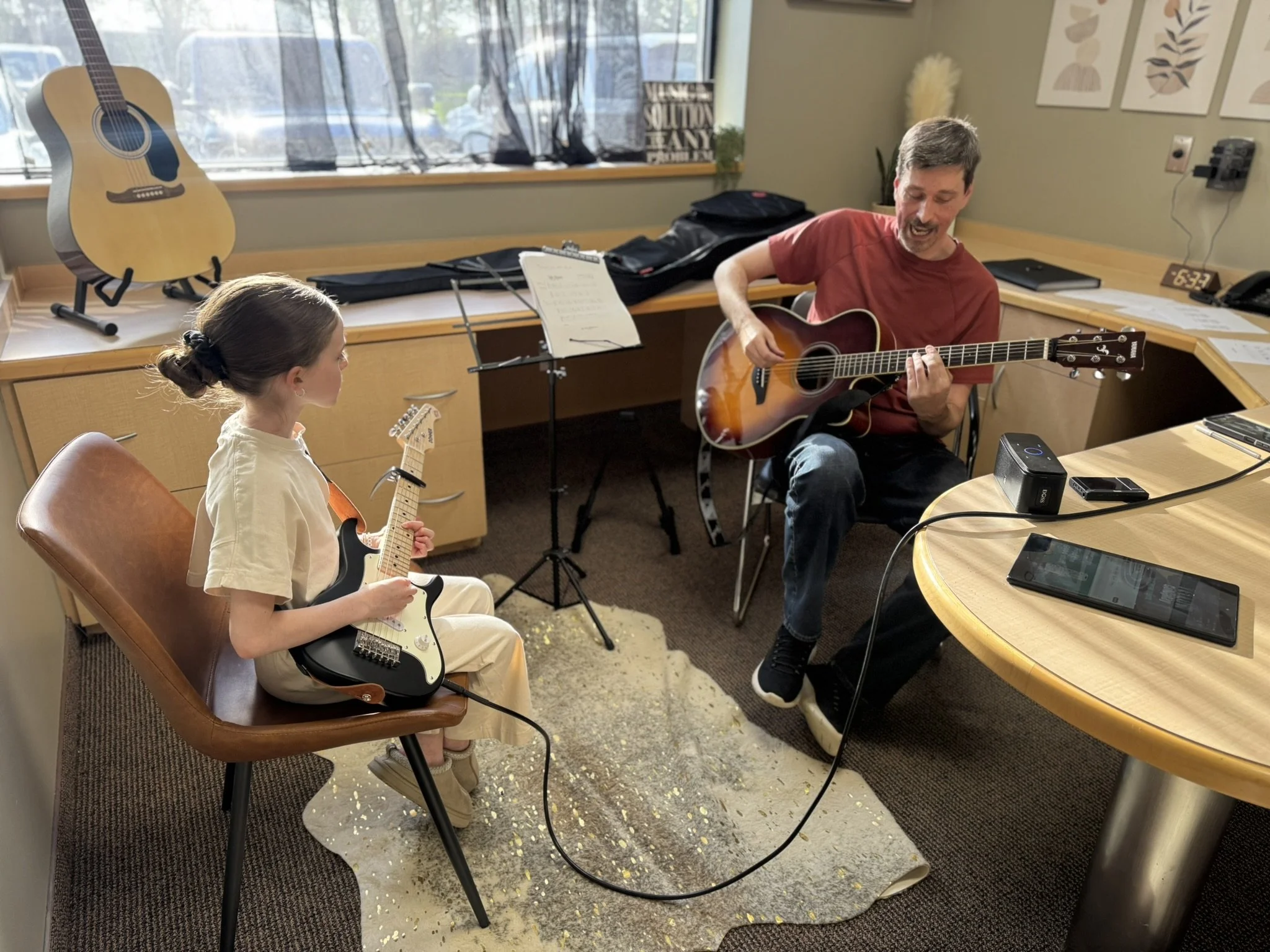 A man and a young girl playing guitars in an indoor room with a large window, artwork on the wall, and various music equipment and papers on the table.