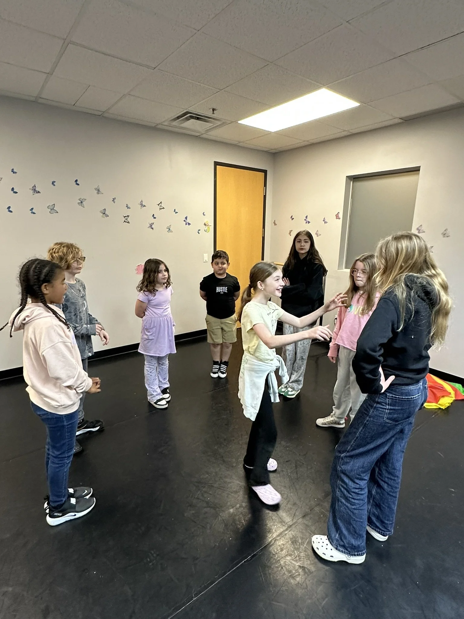 A group of children standing in a circle in a room with butterfly decorations on the wall, some wearing casual clothes, during a group activity or game.