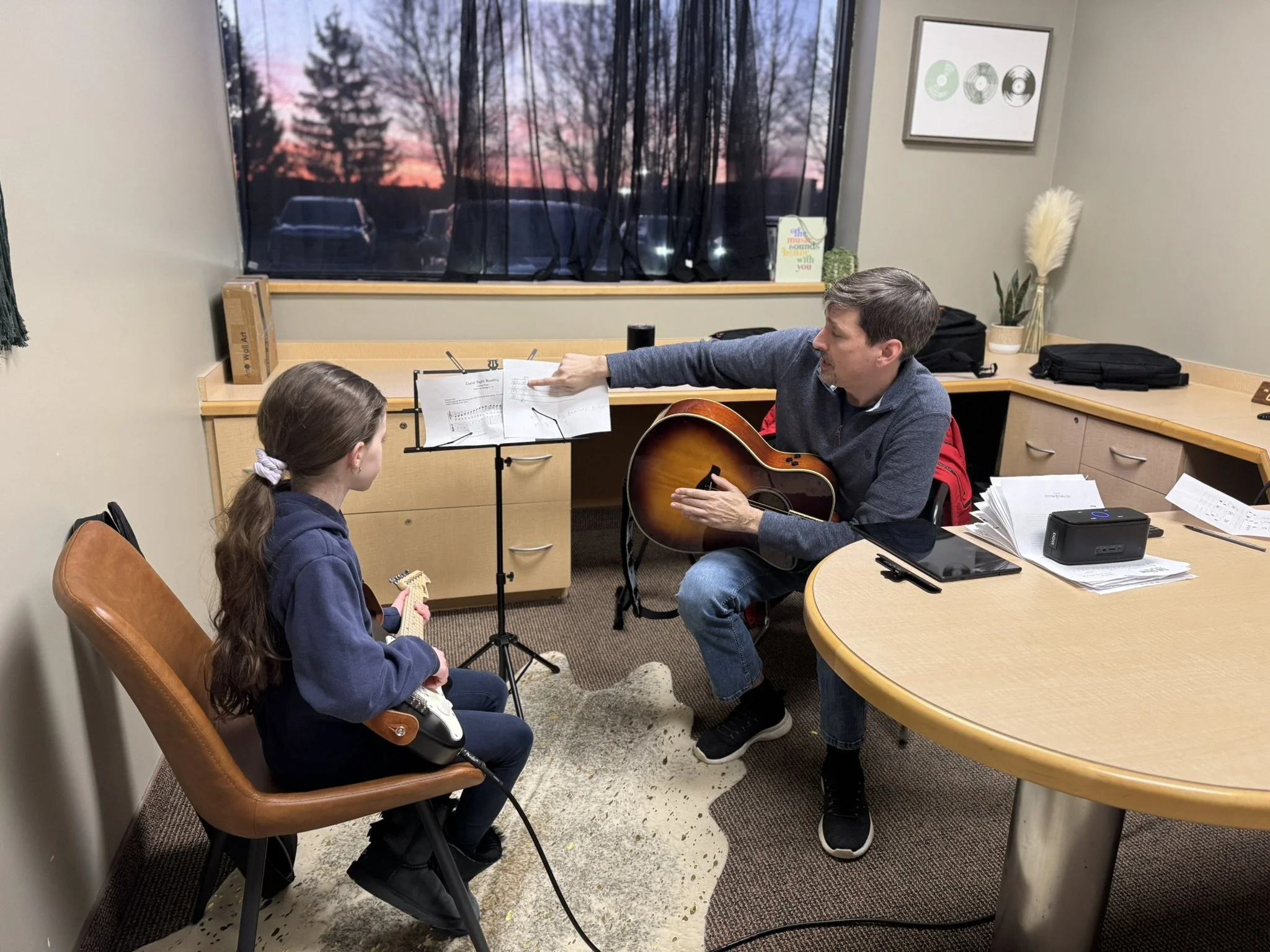 A man playing guitar and singing to a girl in a room with a window showing trees and a sunset sky, with music sheets, a music stand, and various items on the desk.
