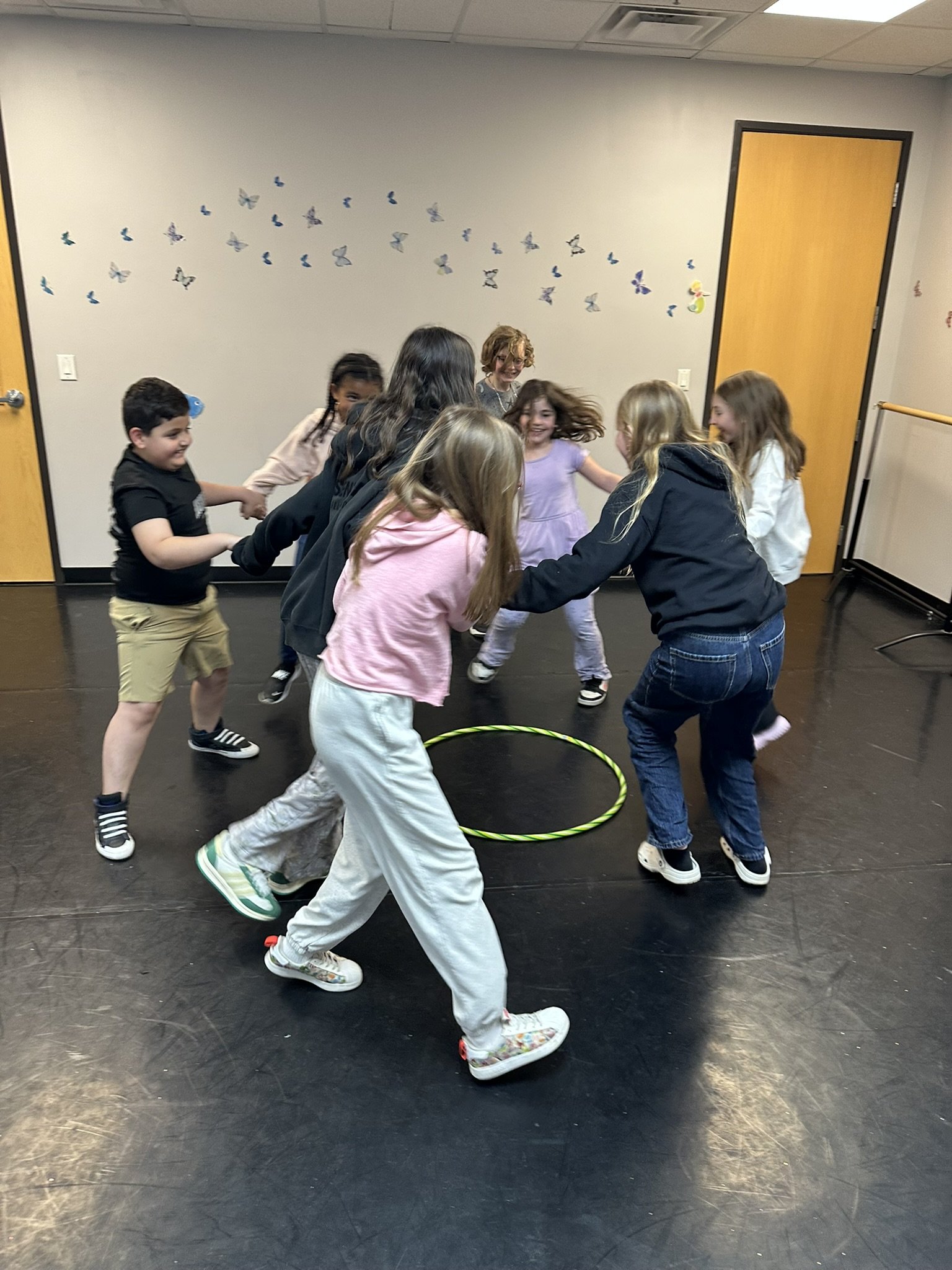 Group of children holding hands and playing in a circle indoors, with butterfly decorations on the wall behind them.