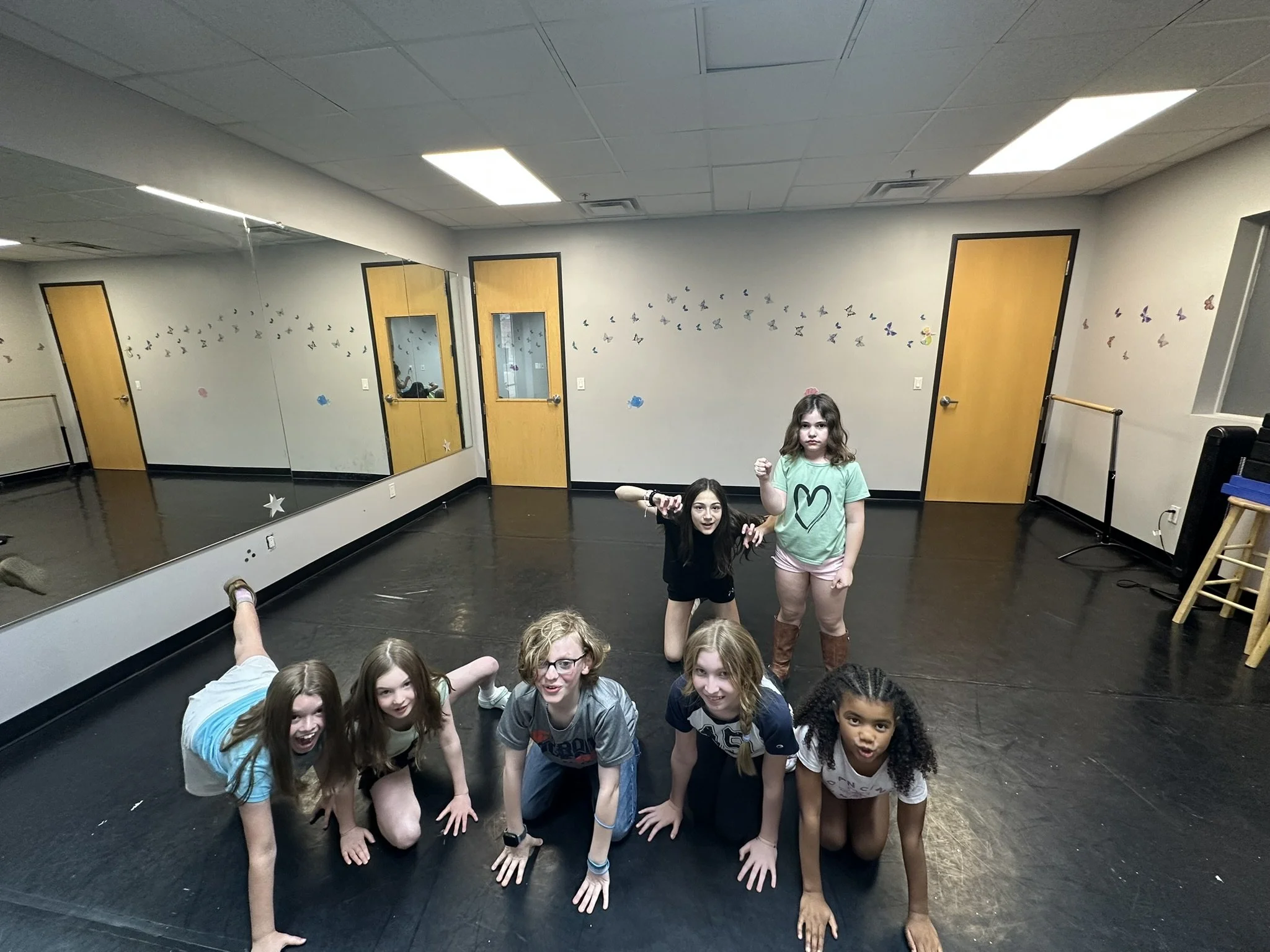 Group of children in a dance studio, some standing and some crouching, with butterfly decorations on the wall and mirrors reflecting the room.