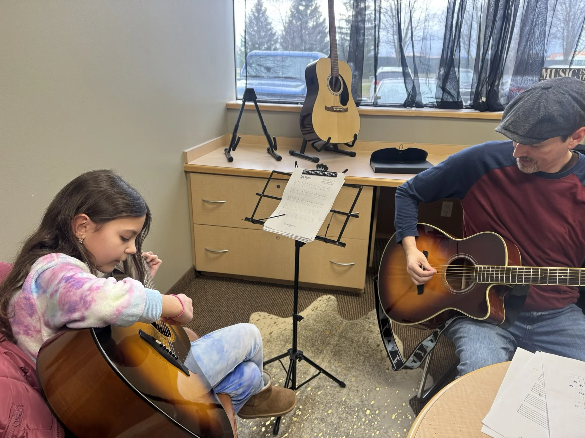 A young girl and a man are sitting in a room with guitars, music stand, and sheet music. The girl is holding a guitar, and the man is playing an acoustic guitar. There is a large window behind them with trees and parked cars visible outside.