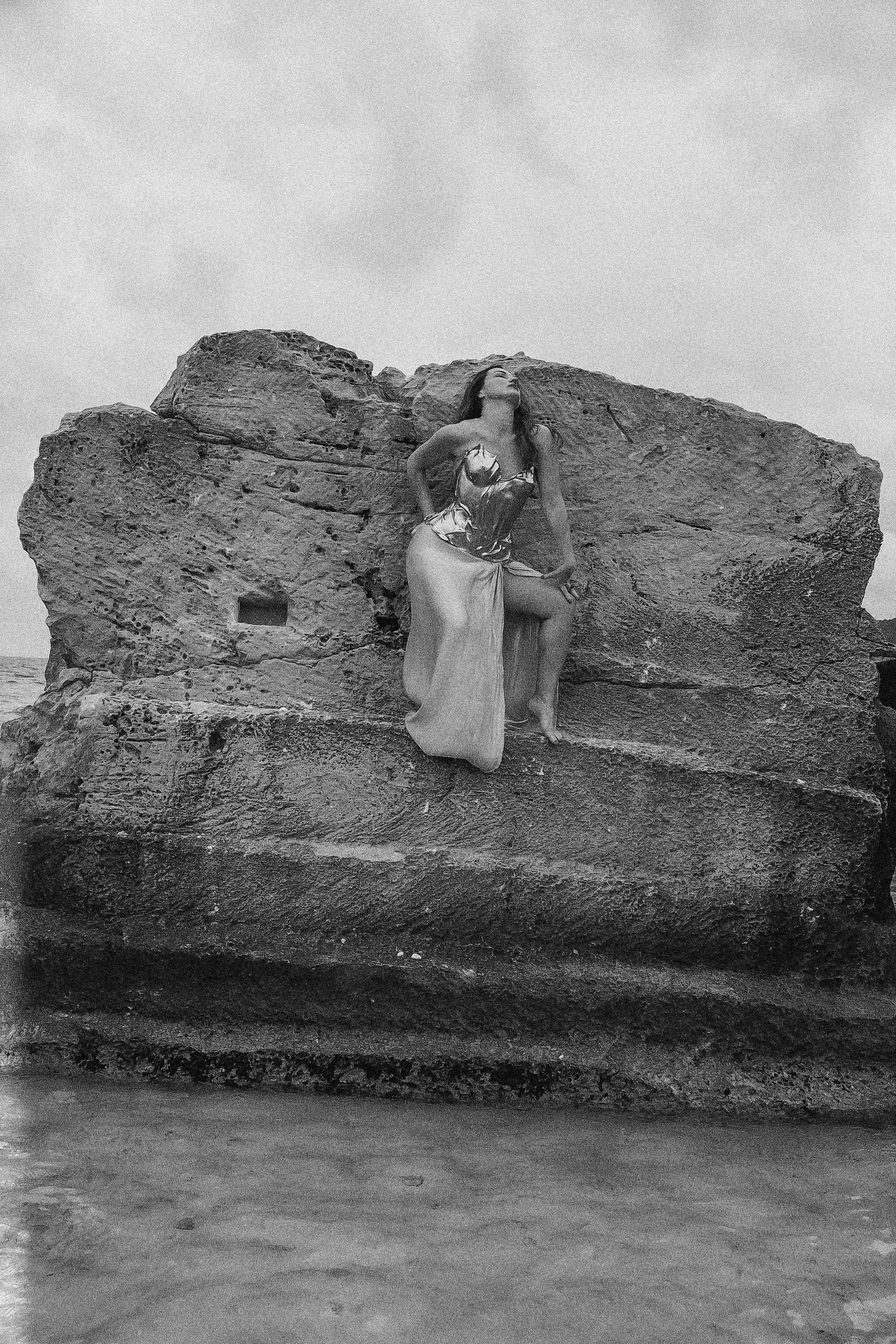 A woman kneels on a large rock formation on a beach, looking upward with her hands on her hips, wearing a dress, with waves and cloudy sky in the background.