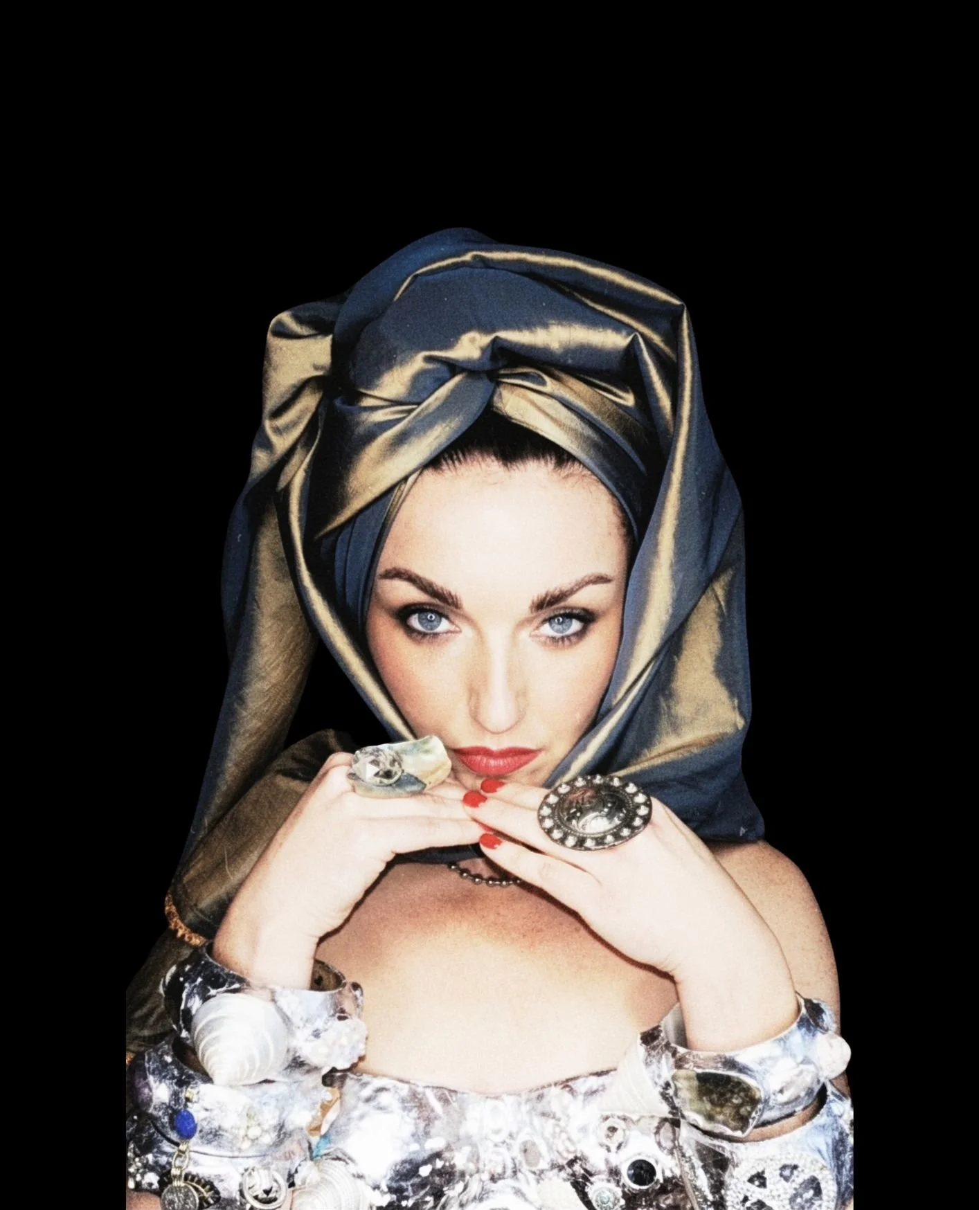A woman with blue eyes and red lipstick, wearing a metallic headscarf, jewelry, and rings, resting her chin on her hands with seashells and jewelry.