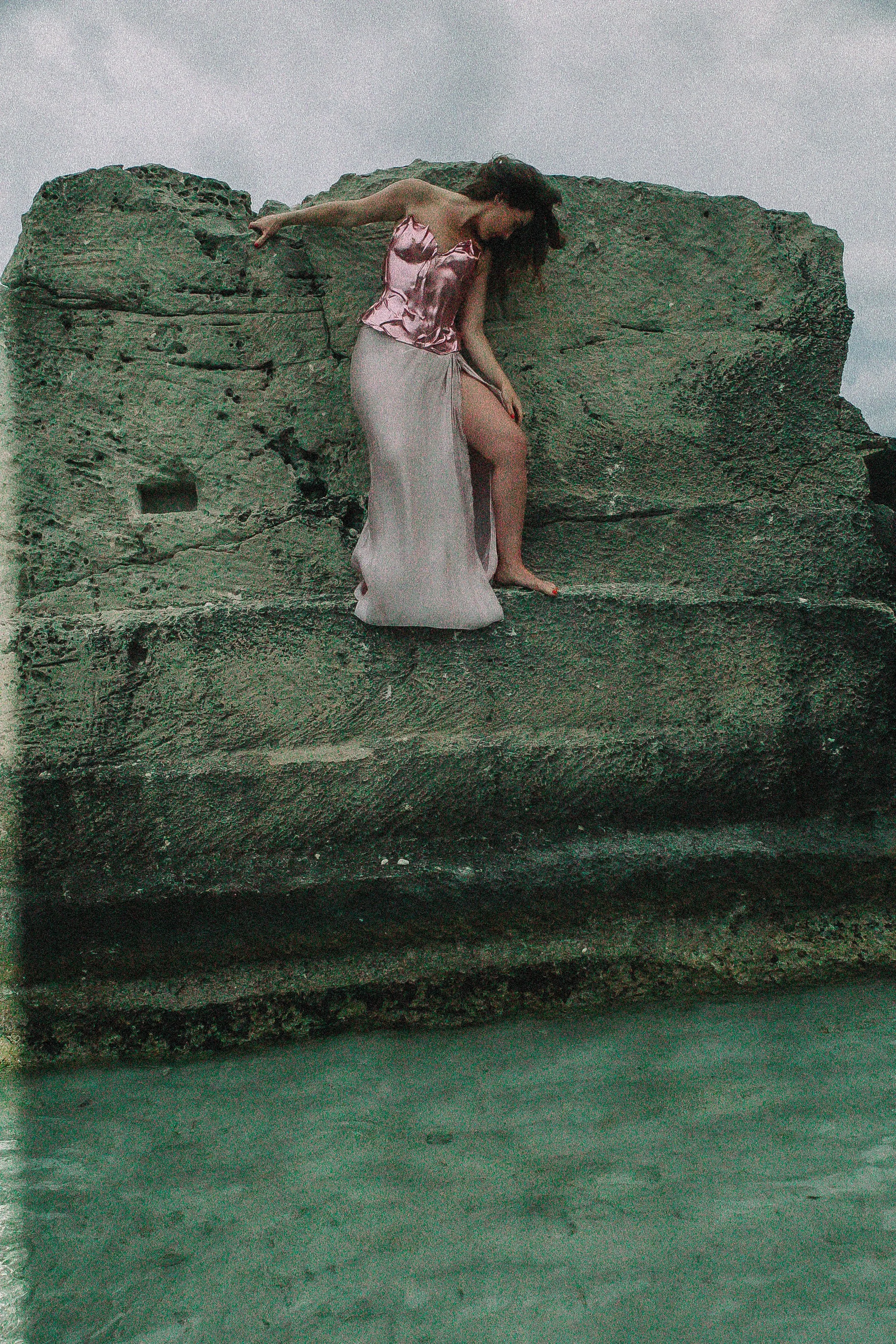 A woman wearing a shiny pink gown standing on a rock formation by the water, with her head and body bent downward and her long dark hair covering her face.