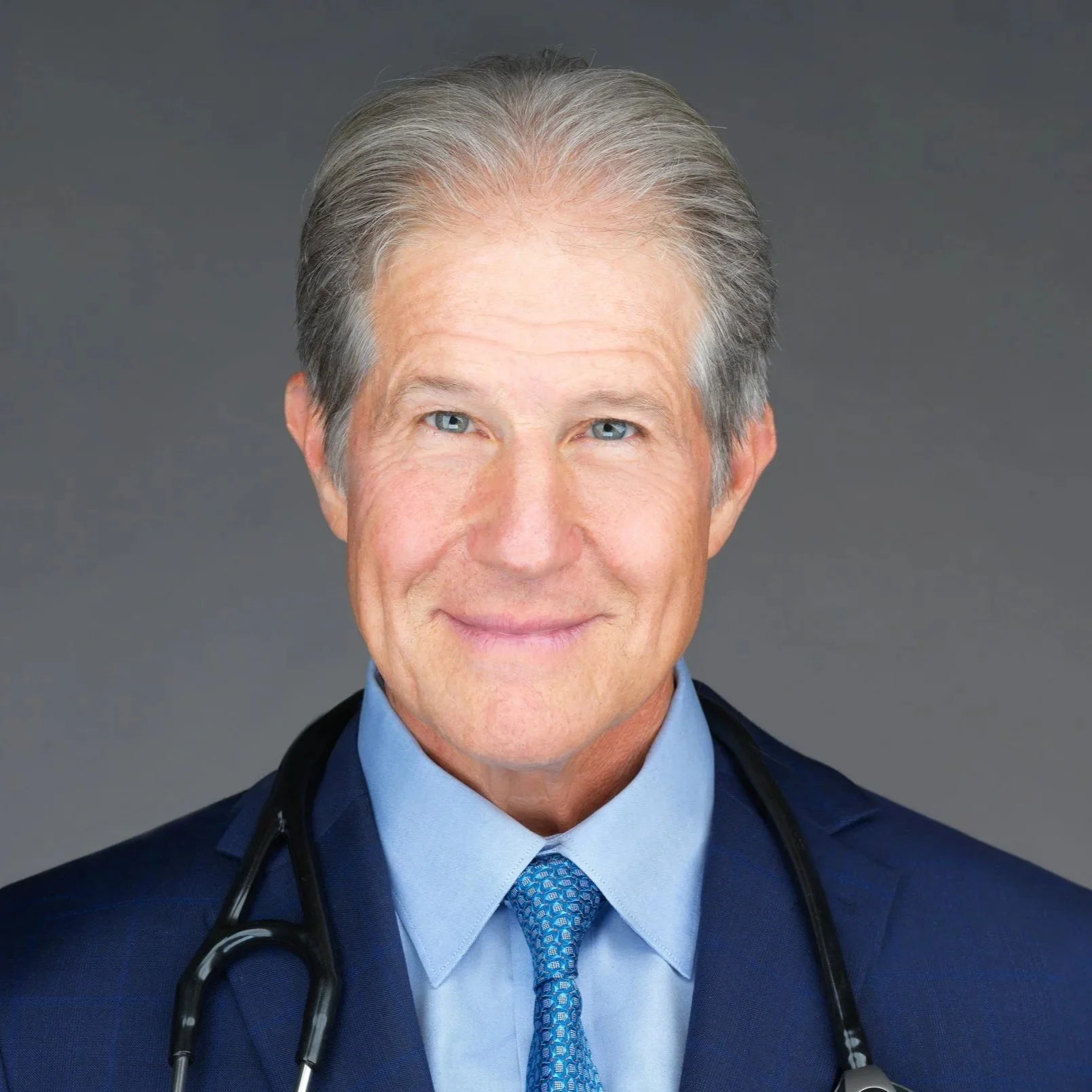 Wayne Jonas, MD with grey hair, wearing a navy blue suit and a light blue shirt, has a stethoscope around his neck and is smiling, against a grey background.