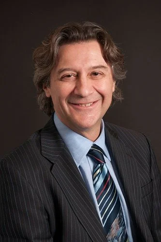 Alessio Fasano, MD wearing a dark pinstripe suit, light blue dress shirt, and a colorful striped tie, smiling against a dark background.