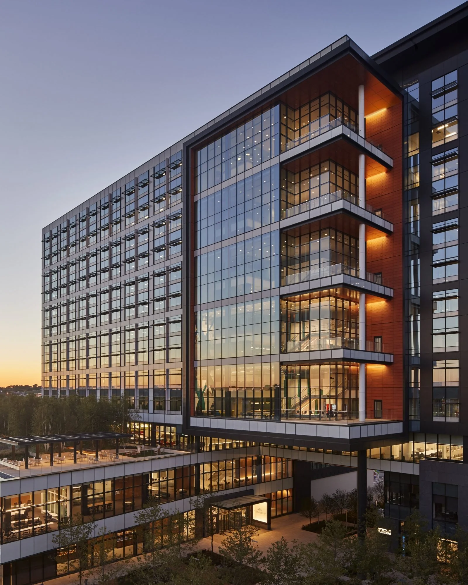 Modern multi-story office building with glass windows and balconies, illuminated from within, during sunset.