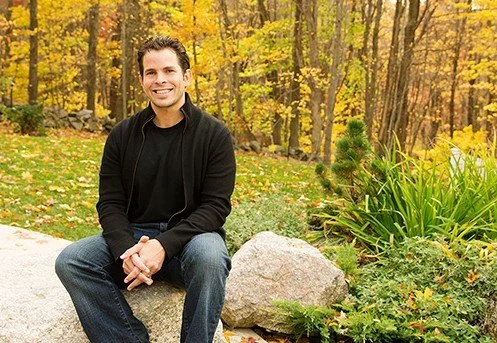 Presenter, Nick Ortner, seated on rock in front of wooded yard or park