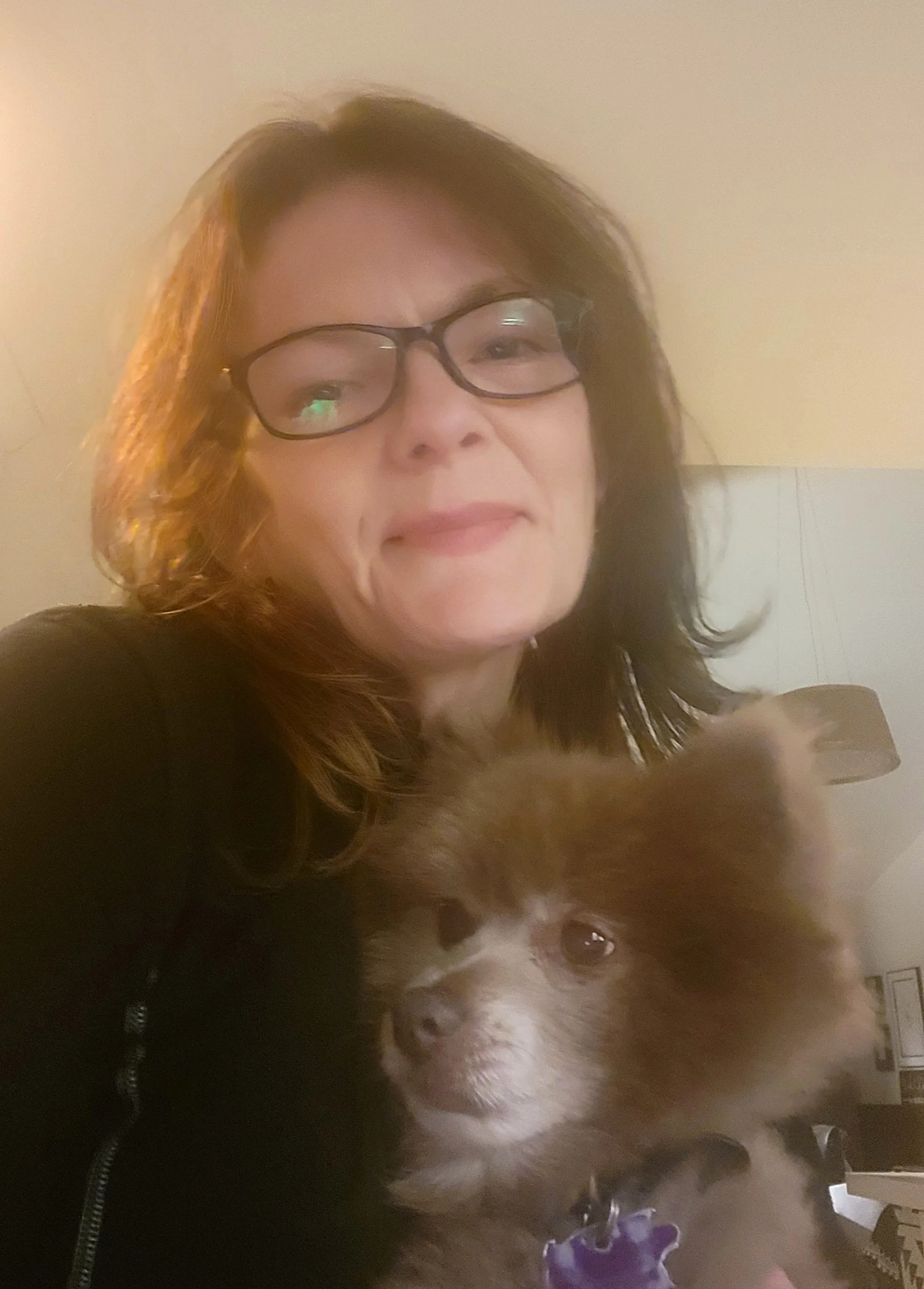 A woman with glasses and dark hair smiling, holding a dog with a white and brown coat and purple collar, indoors against a beige wall.