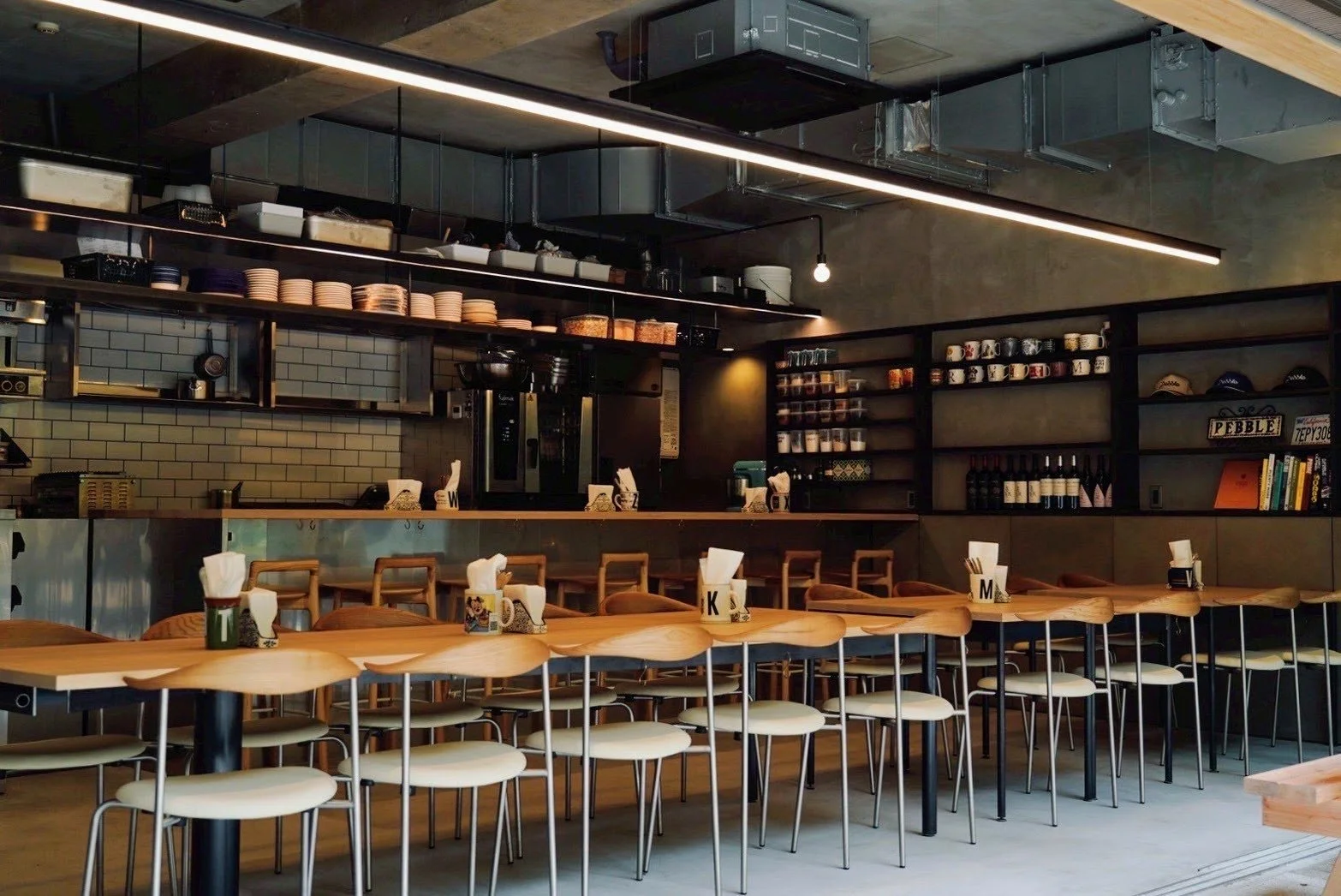 Interior of a modern cafe with a long wooden dining table, chairs, and a bar area with shelves of cups and bottles, and espresso machine in the background.