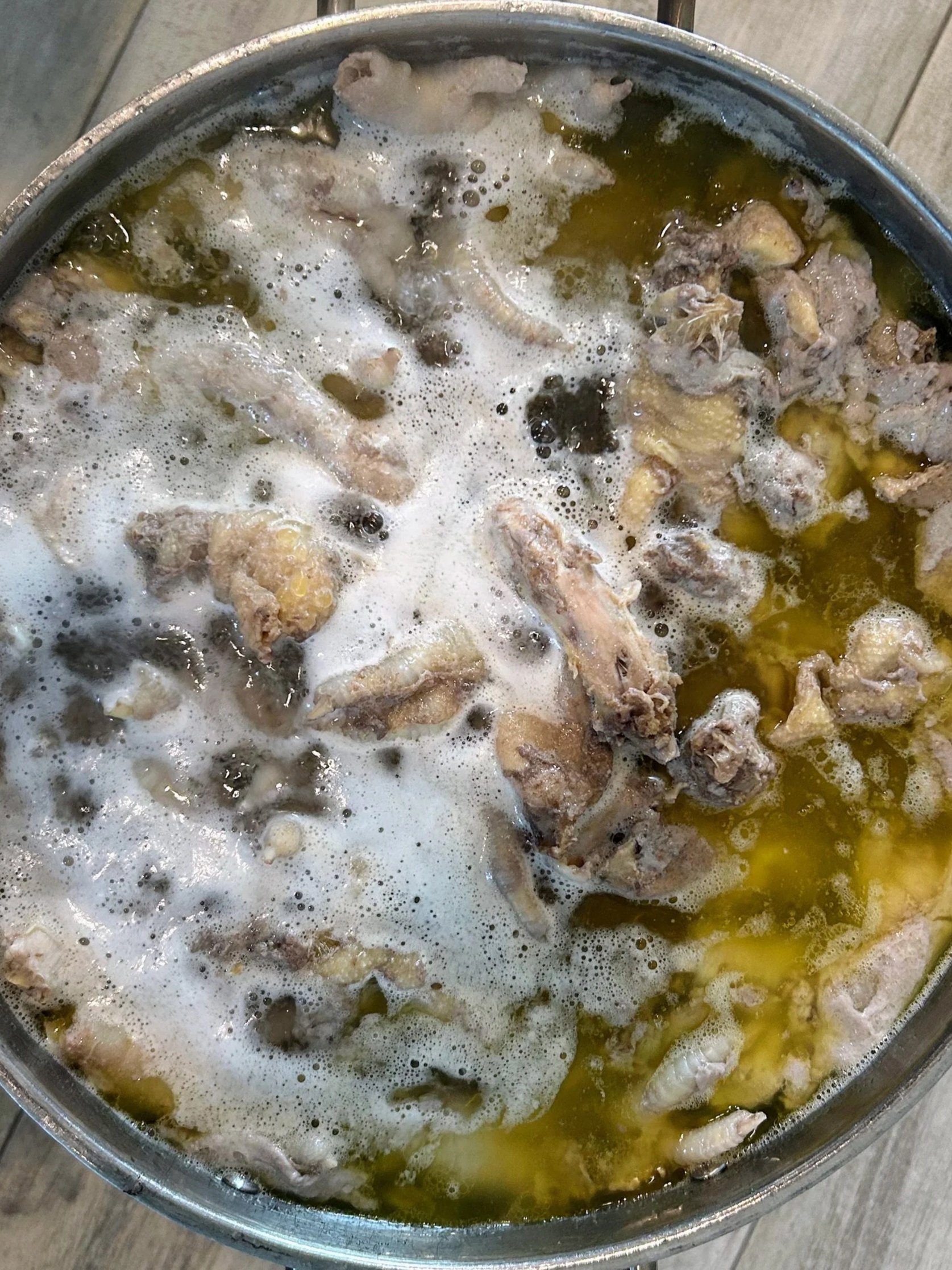 A metal bowl filled with boiling broth containing chicken pieces and fat.