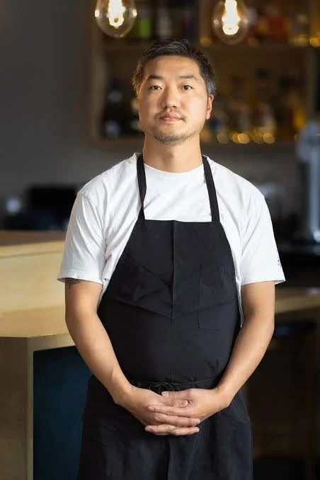 Man standing in a kitchen wearing a white t-shirt and black apron, with shelves and bottles in the background.