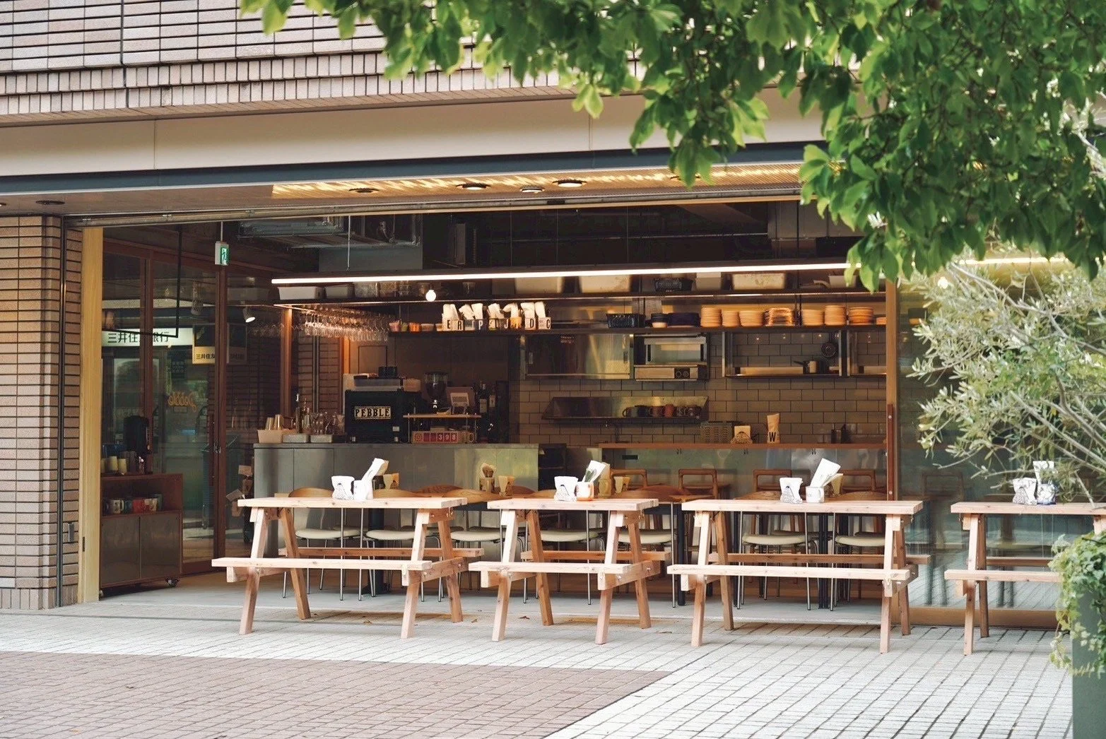 Empty outdoor seating area with wooden tables and chairs outside a modern cafe, with large glass windows revealing the interior counter and kitchen.