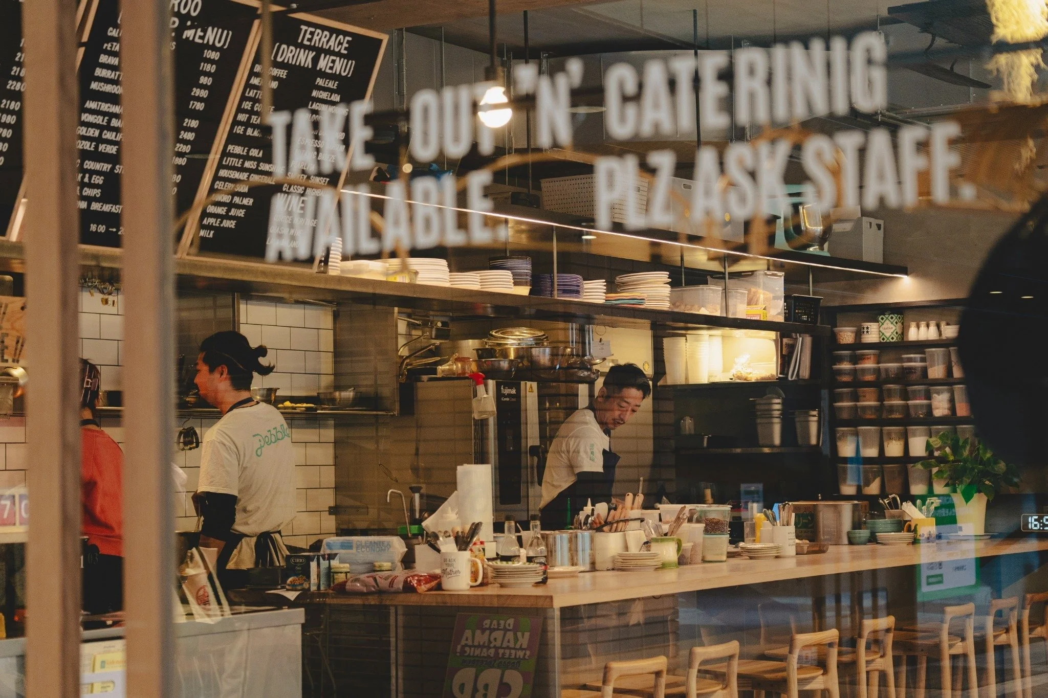 Inside a cafe or restaurant with a counter and menu boards. Two staff members working behind the counter, with one smiling. Various kitchen equipment, plates, and utensils are visible on the counter and shelves. The scene is viewed through a window or glass partition.