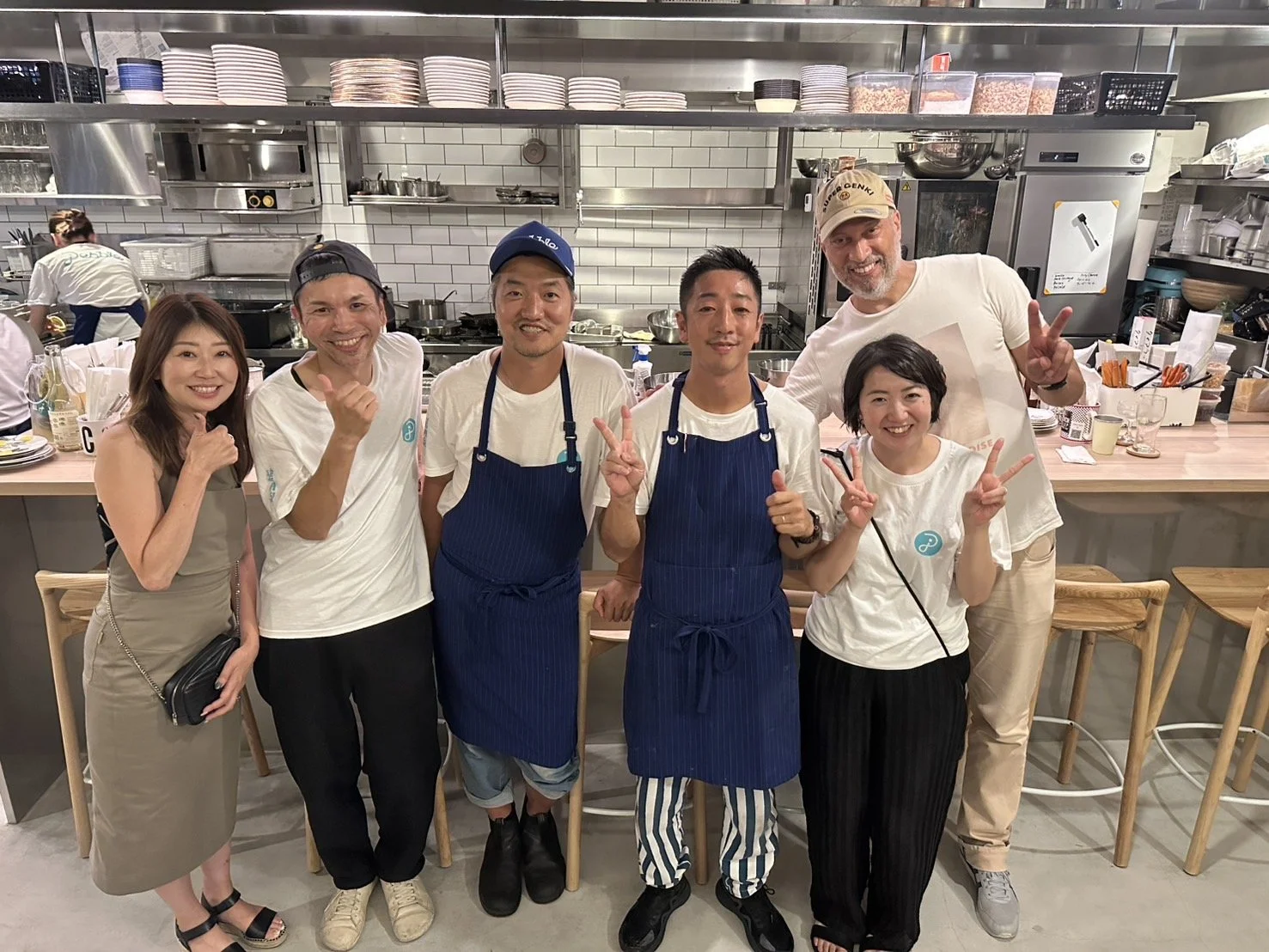 Group of seven people, including three chefs in blue aprons and four customers, smiling and posing inside a restaurant kitchen.