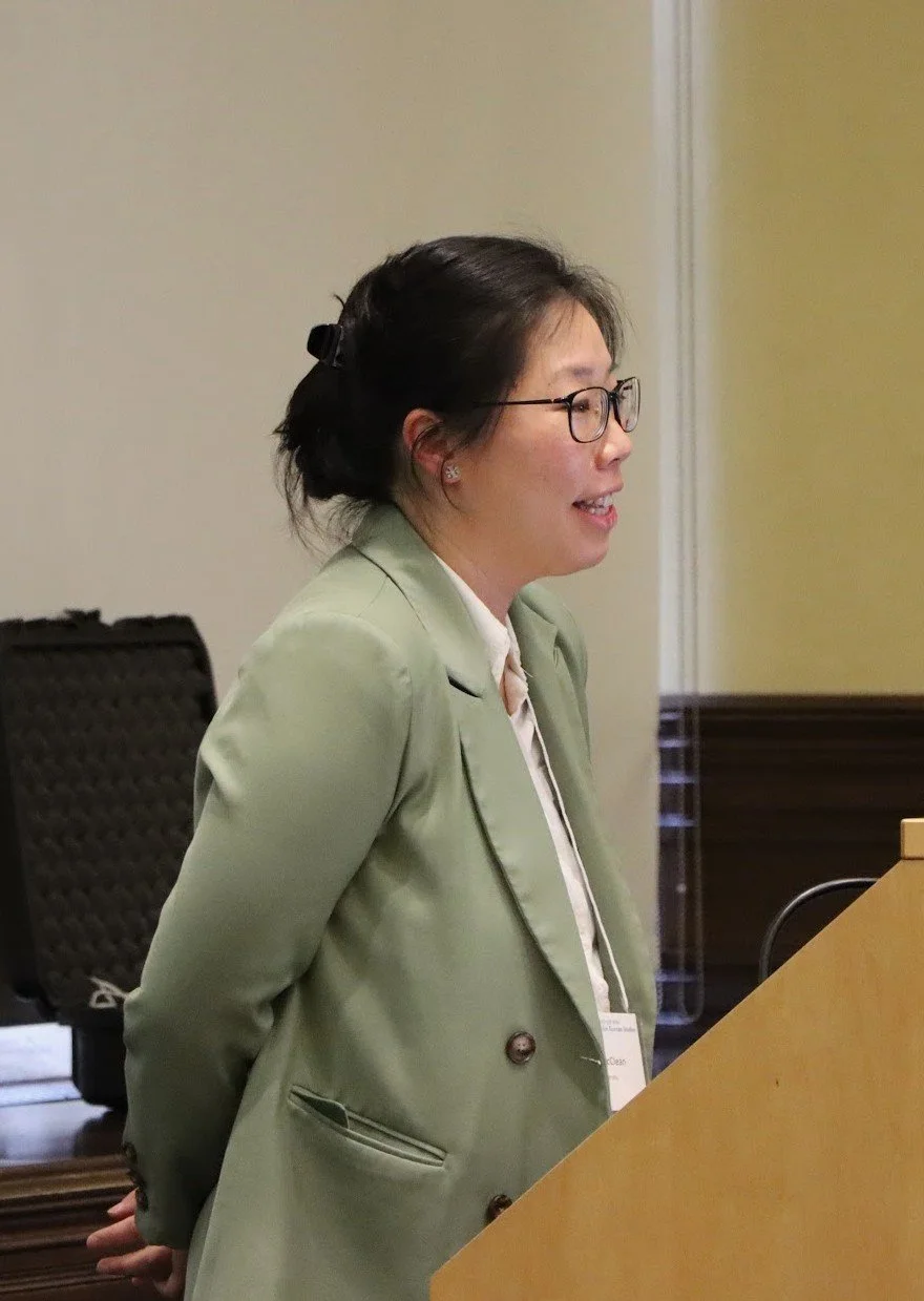 A woman wearing glasses and a light green blazer standing behind a podium in a conference room.