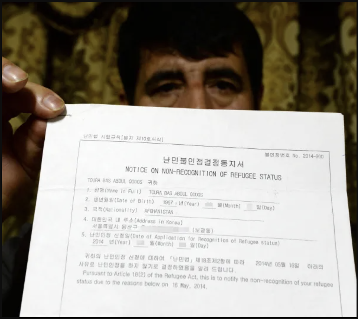 Man holding a document titled 'Notice on Non-Recognition of Refugee Status' with Korean and English text, in front of a patterned curtain.