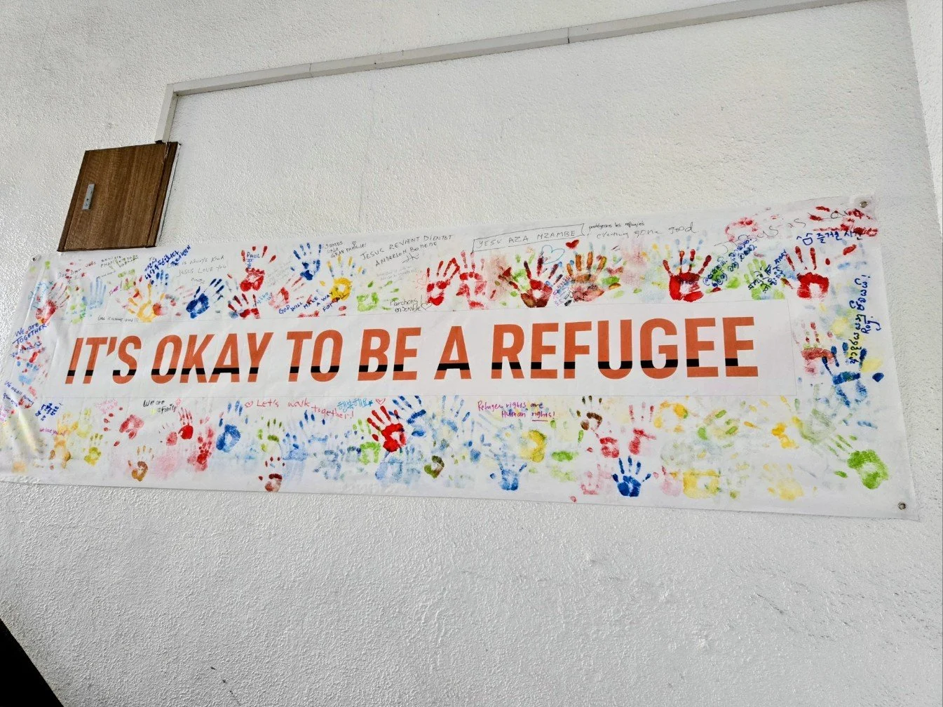 A large white banner on a wall that says, 'IT'S OKAY TO BE A REFUGEE' in bold orange and black letters. The banner is decorated with colorful handprints and handwritten messages around the border.